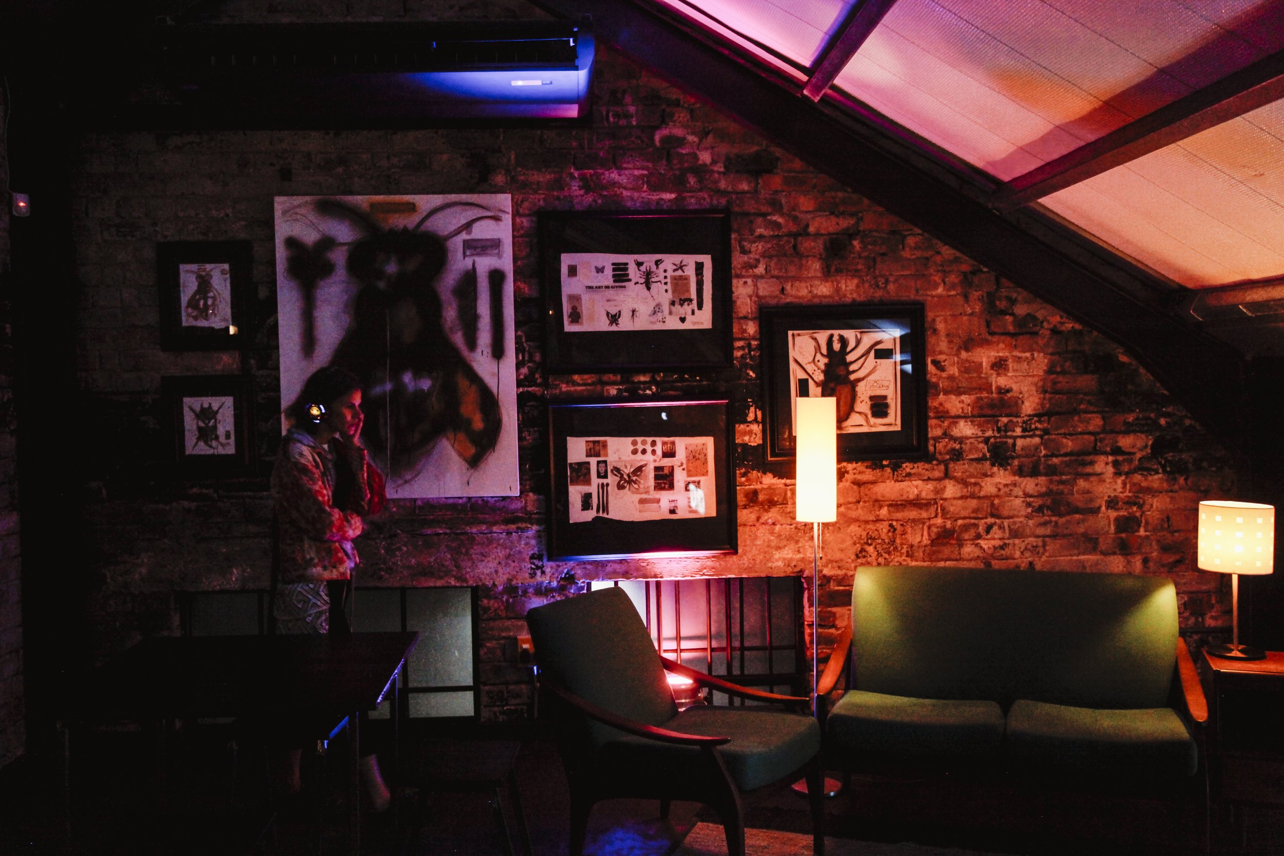 A dimly lit lounge with brick walls, artwork of insects, and a woman in headphones standing near a table.