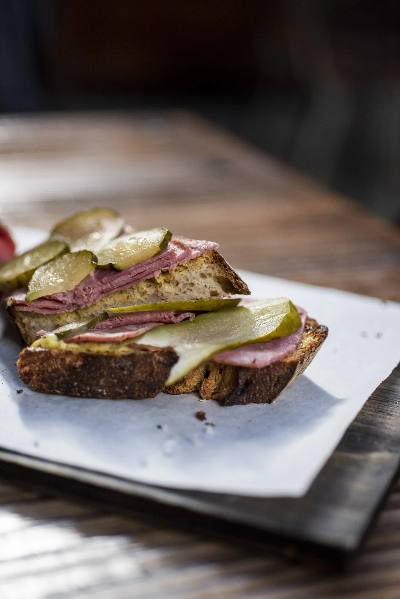 Close-up of a sandwich with pickles and meats on toasted bread, on a piece of white paper on a wooden surface.