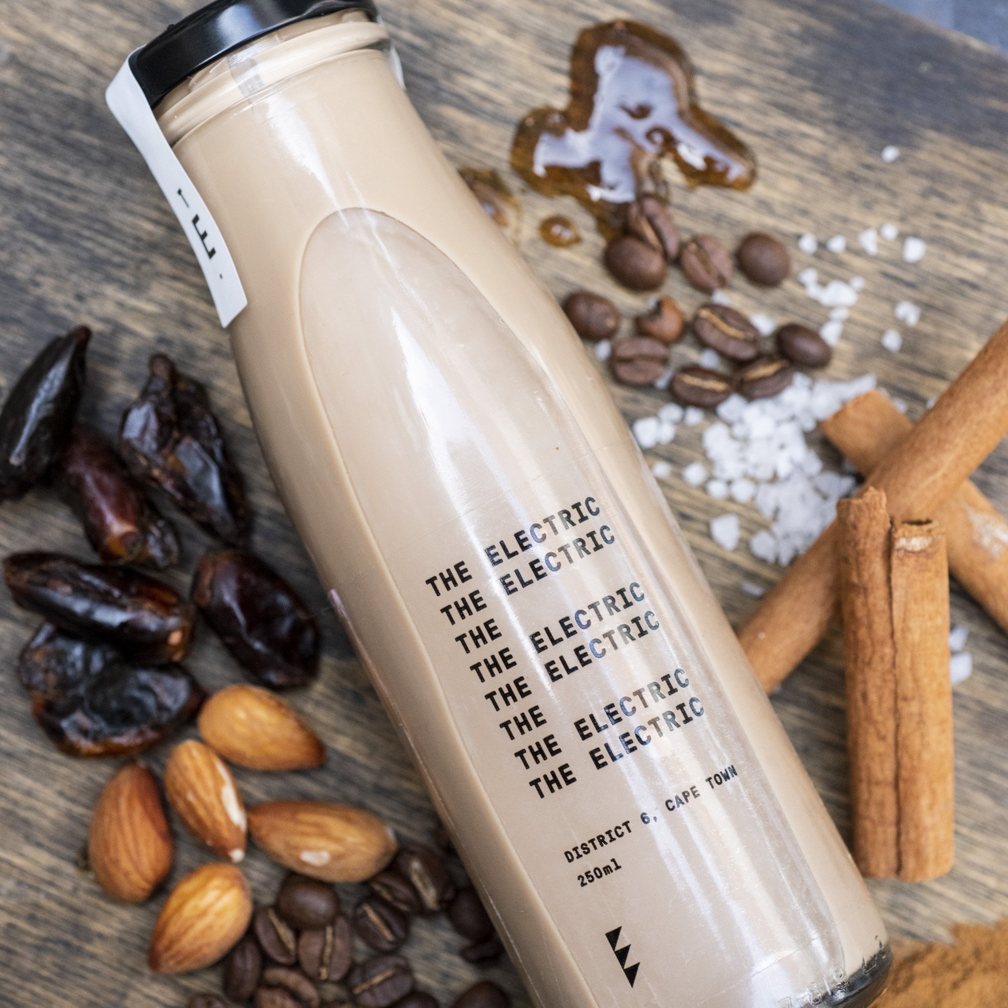A bottle of coffee-flavored beverage labeled "The Electric" surrounded by date fruit, almonds, coffee beans, salt, and cinnamon sticks on a wooden surface.