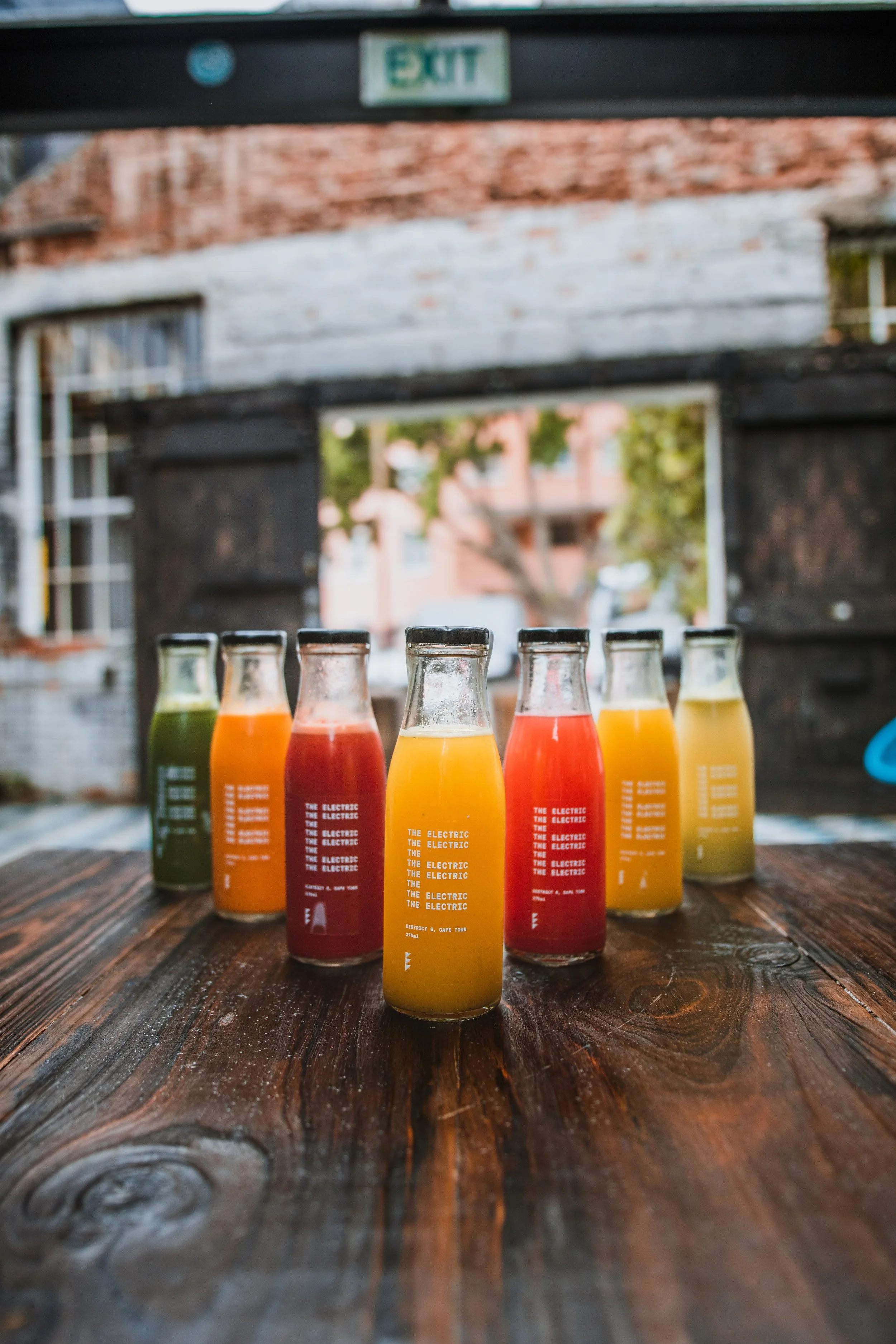 Six bottles of colorful juice on a wooden table outdoors, with a brick and wood structure and an exit sign in the background.