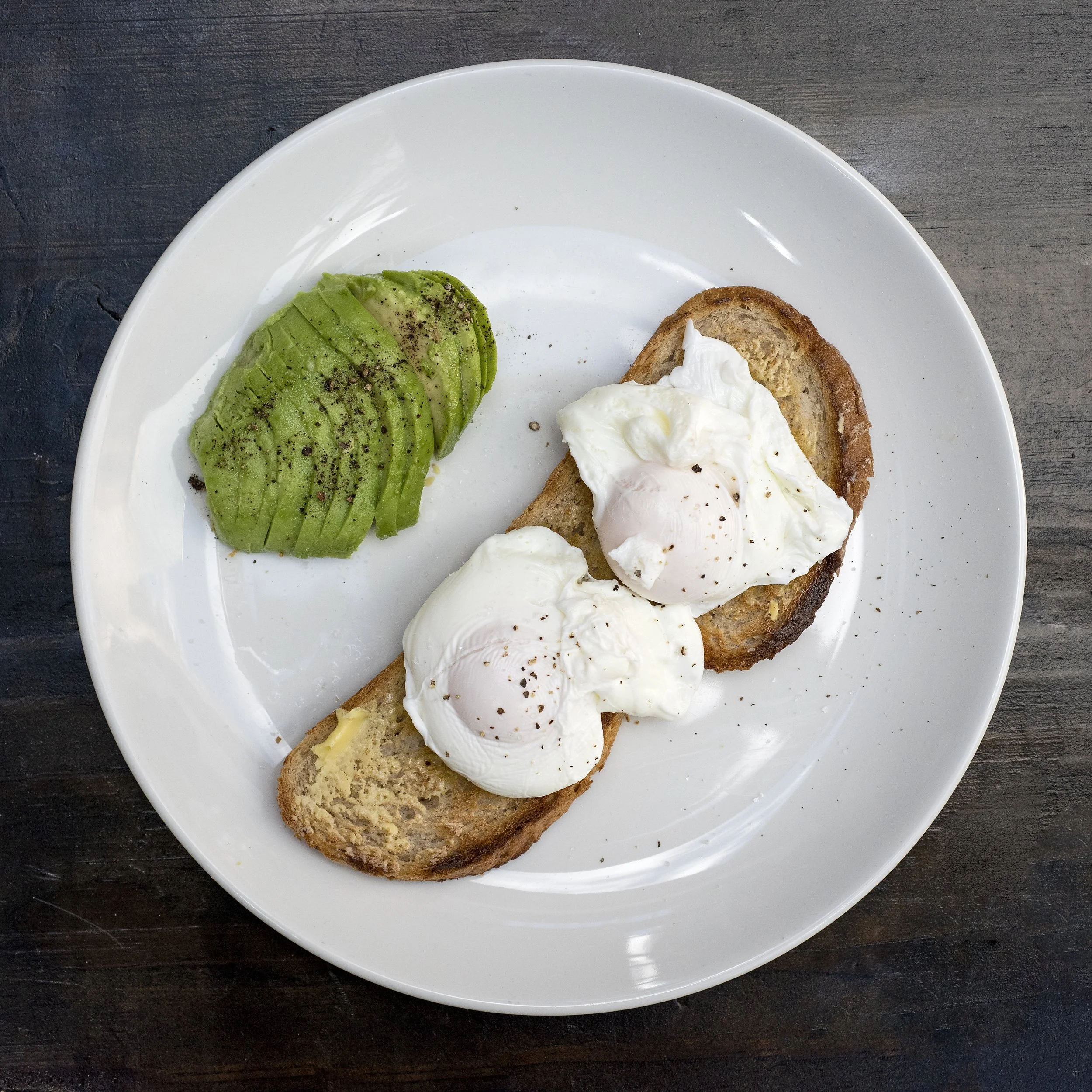 Sliced avocado sprinkled with black pepper and two poached eggs on toasted bread on a white plate.
