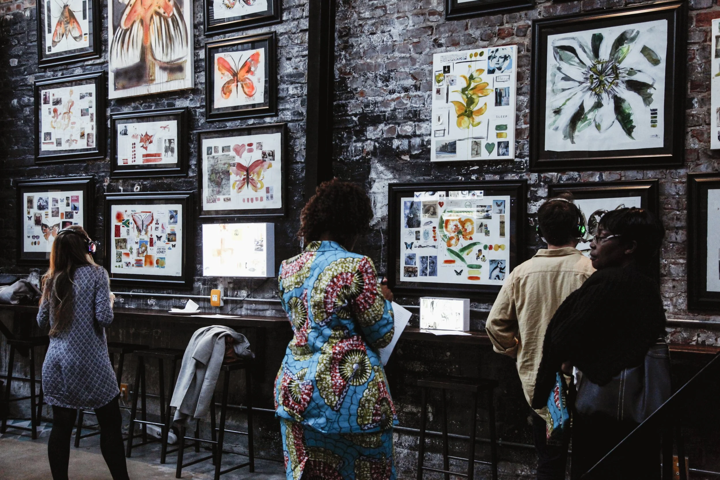 People viewing framed butterfly art and floral prints in an art gallery with a brick wall.