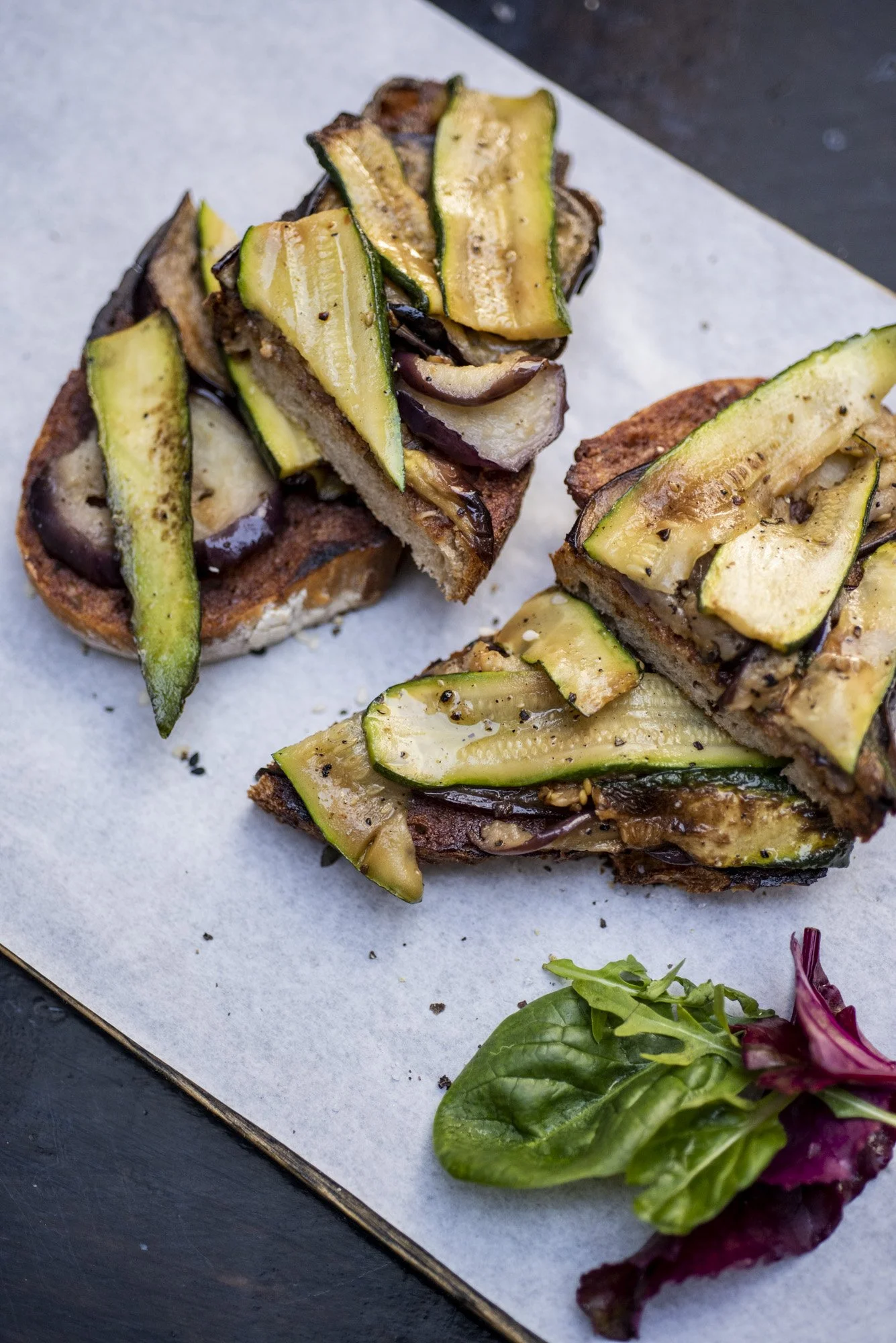 Two pieces of toast topped with grilled zucchini, eggplant, and possibly mushrooms, served on parchment paper with a small side salad of mixed greens.