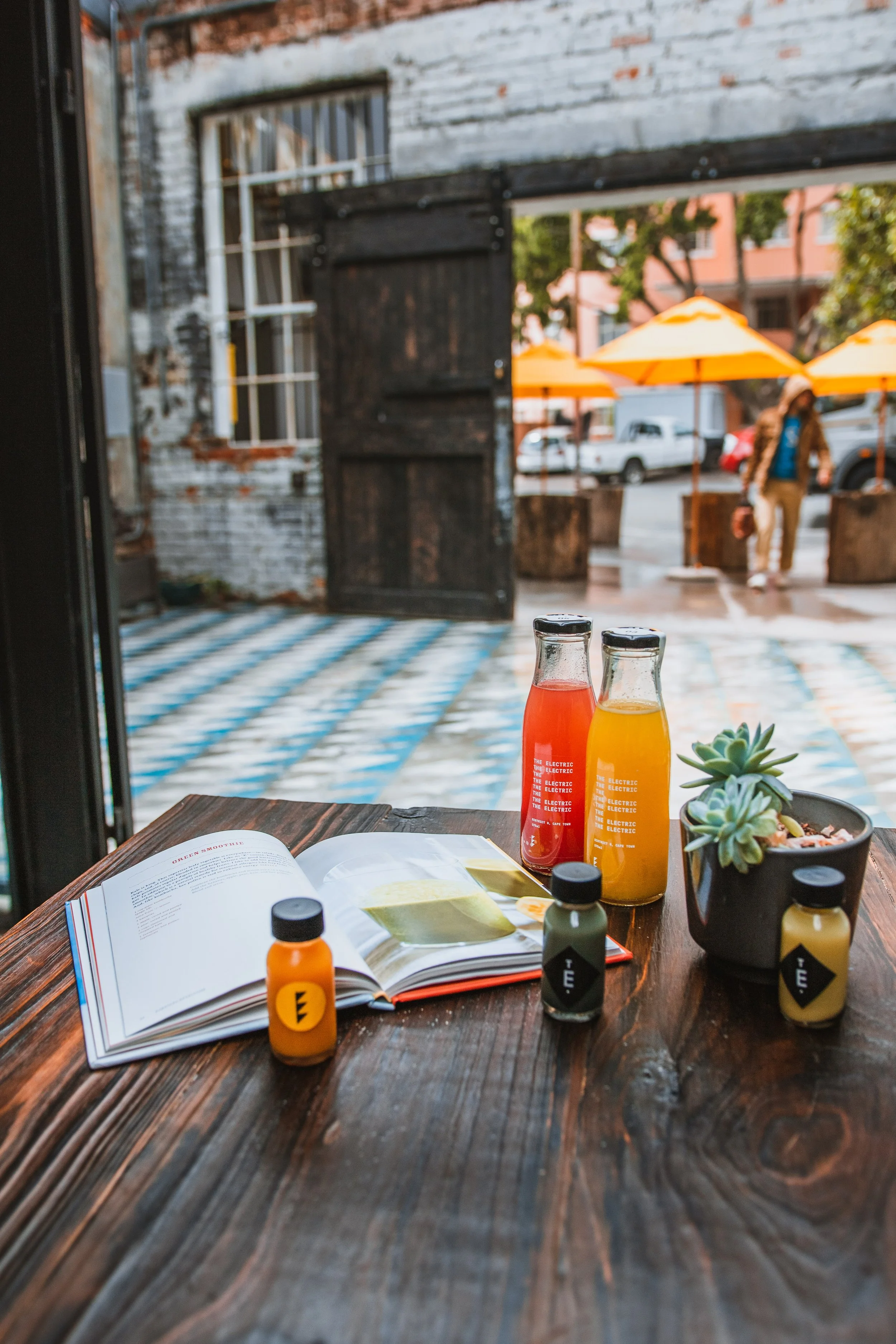 Table with open magazine, three small bottles, two bottles of juice, and a potted succulent plant inside a cafe with a view of outdoor seating area with orange umbrellas and a person walking by.