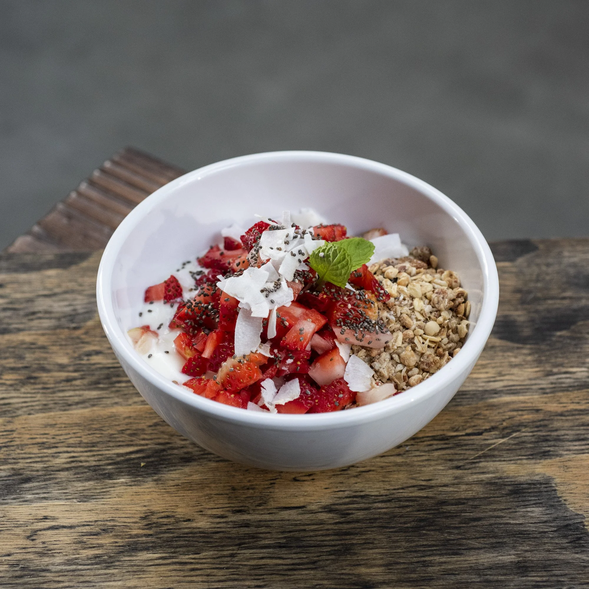 A white bowl of yogurt topped with chopped strawberries, shredded coconut, granola, chia seeds, and a mint leaf on a rustic wooden surface.