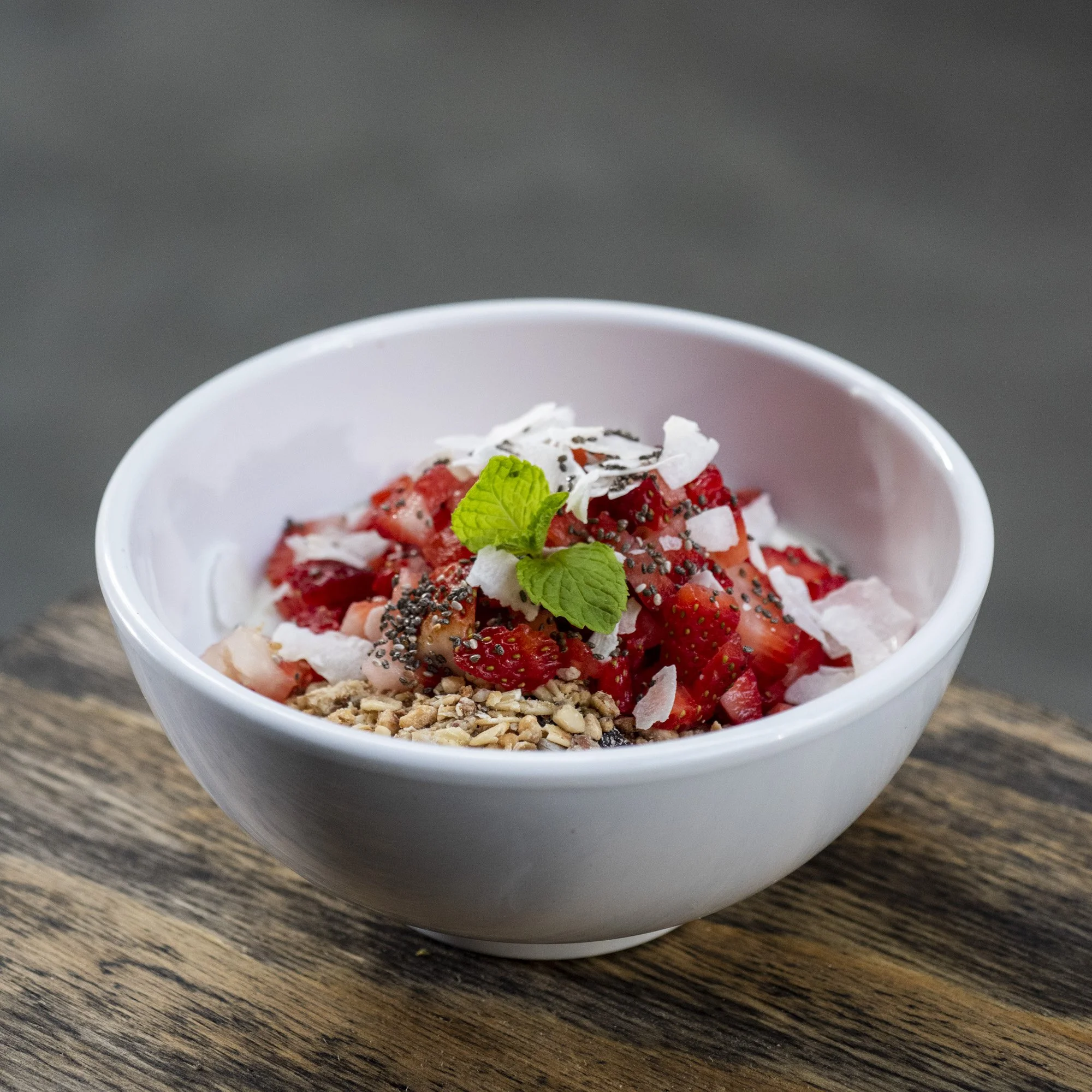 Bowl of mixed strawberries topped with shredded coconut, chia seeds, mint leaves, and chopped nuts on a wooden surface.