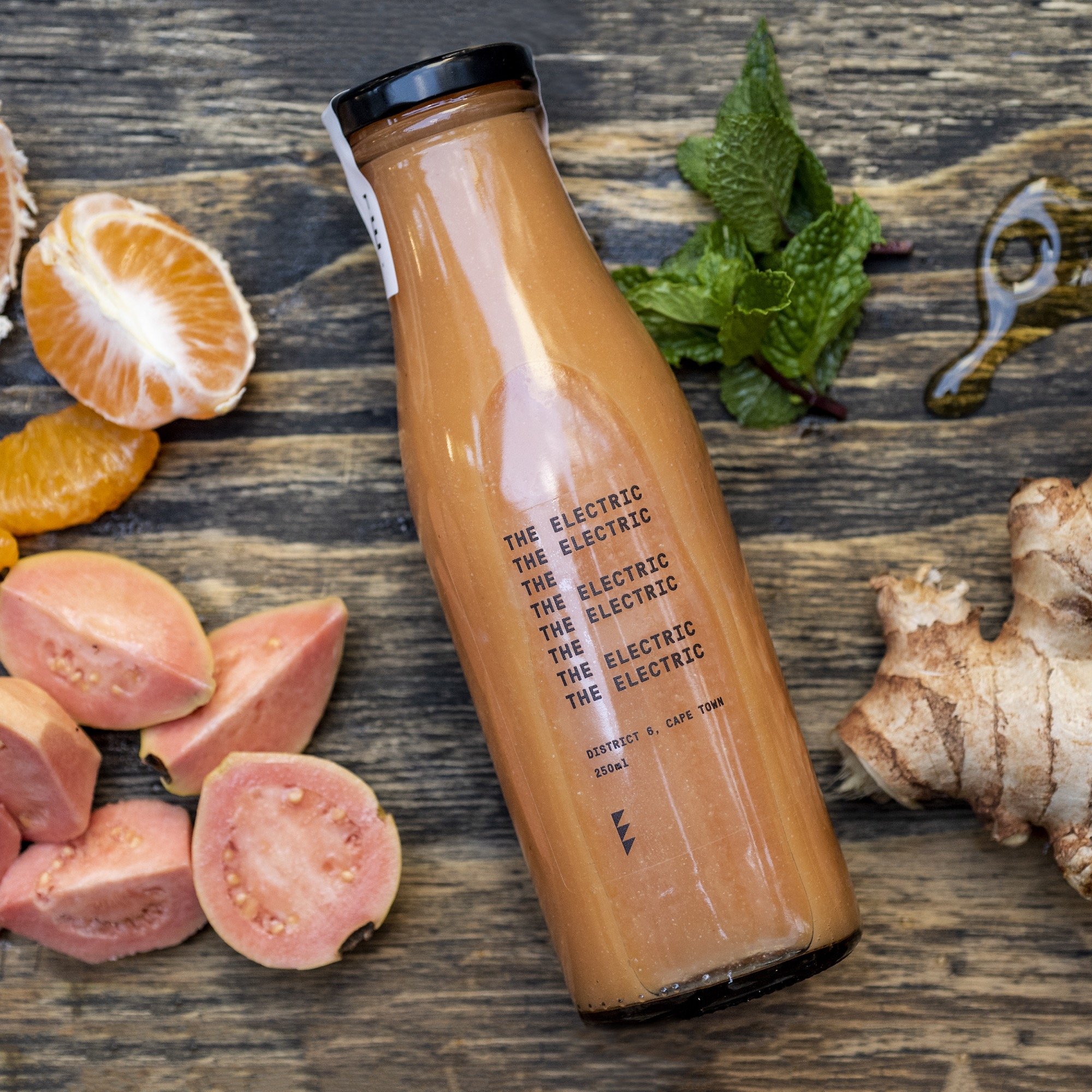 A bottle of orange-colored juice on a rustic wooden surface, surrounded by pieces of pink guava, orange slices, fresh mint leaves, ginger root, and a small amount of honey or syrup.