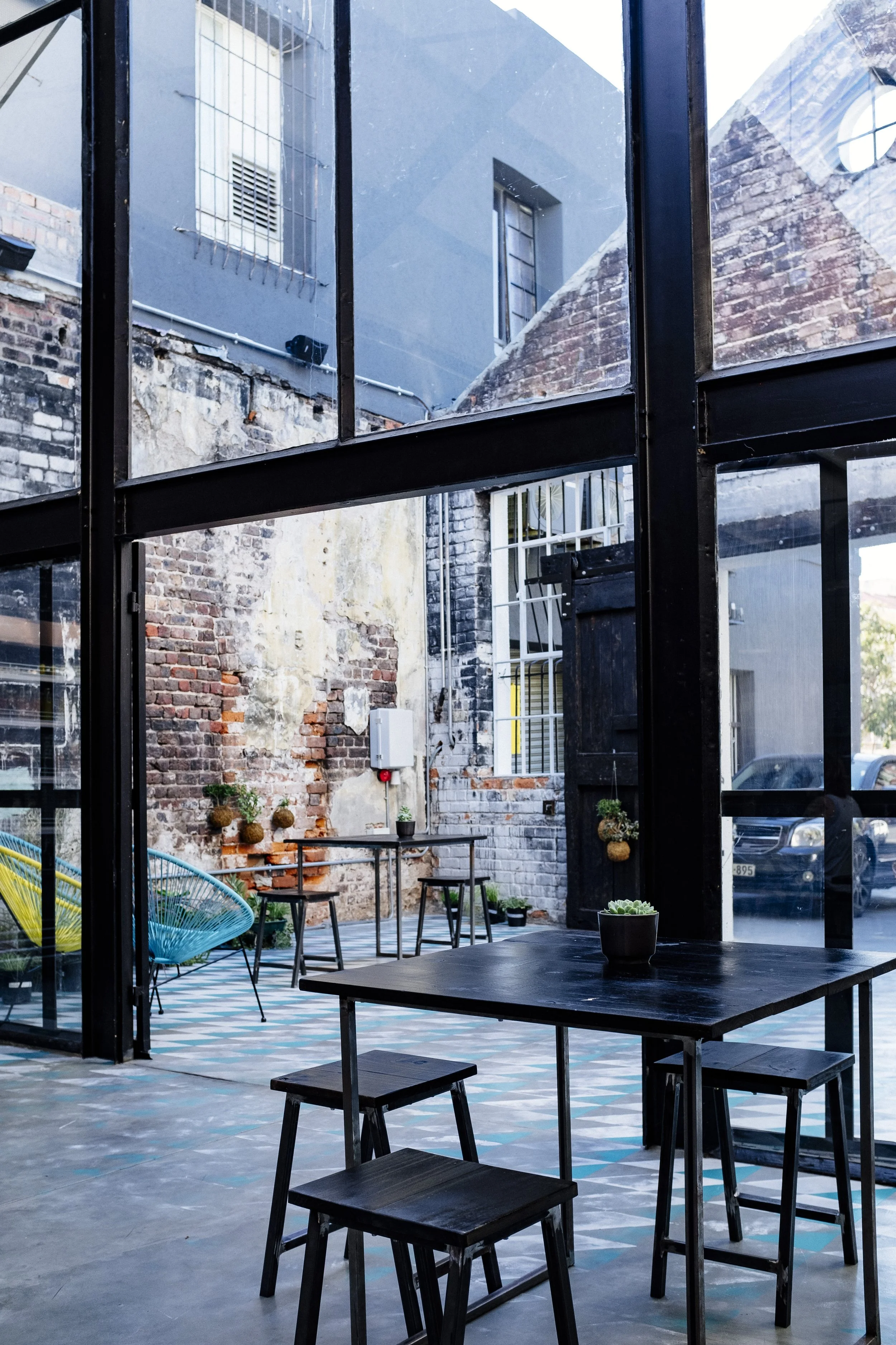 Interior view of a modern, industrial-style cafe with black metal-framed glass walls, exposed brick and concrete walls, small tables, plants, and outdoor seating visible.