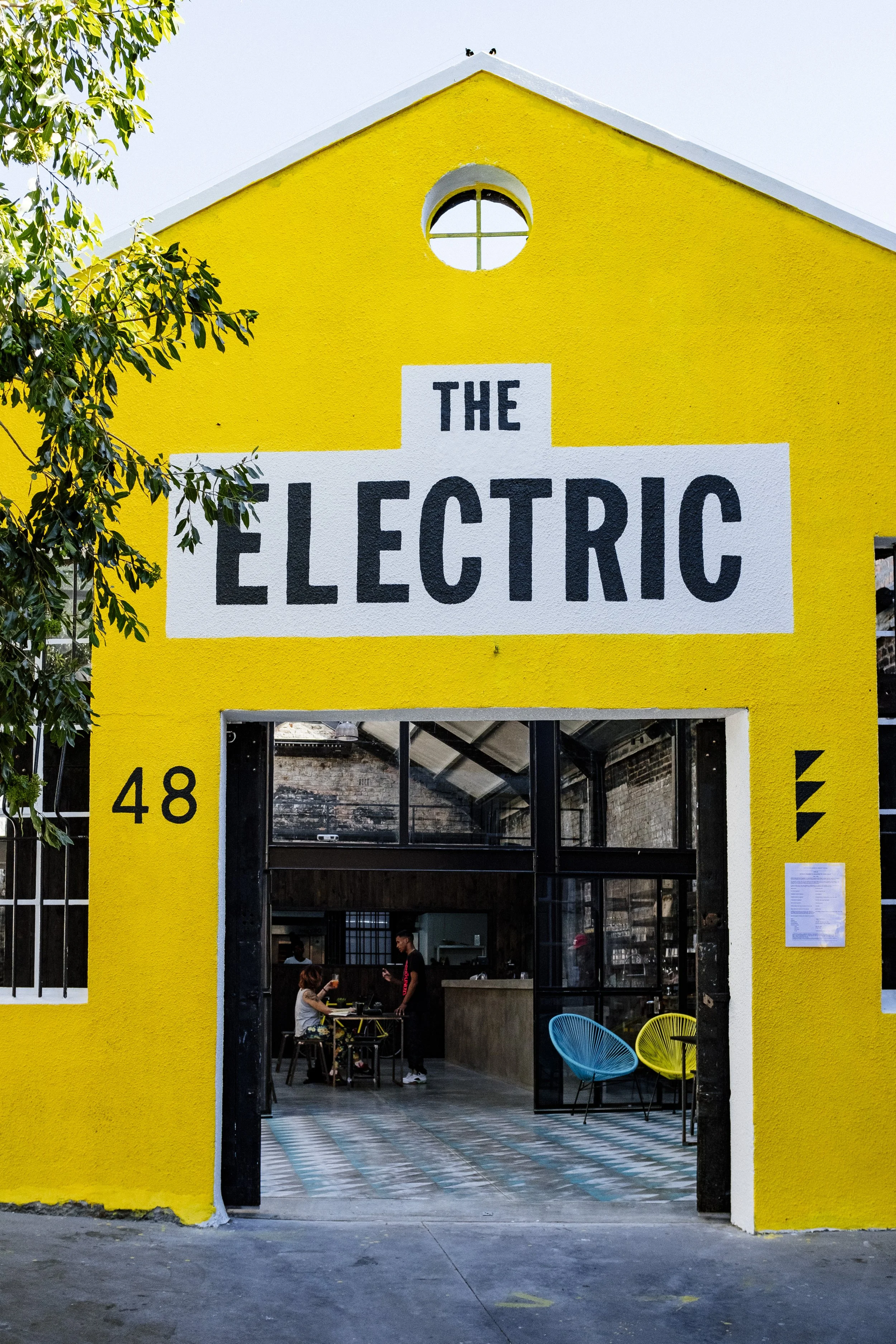 Front view of a yellow building with a sign that says 'The Electric' and the number 48 on the left side of the entrance. The building has large glass doors, and inside there are people sitting at tables, with colorful chairs visible. The weather appears clear.