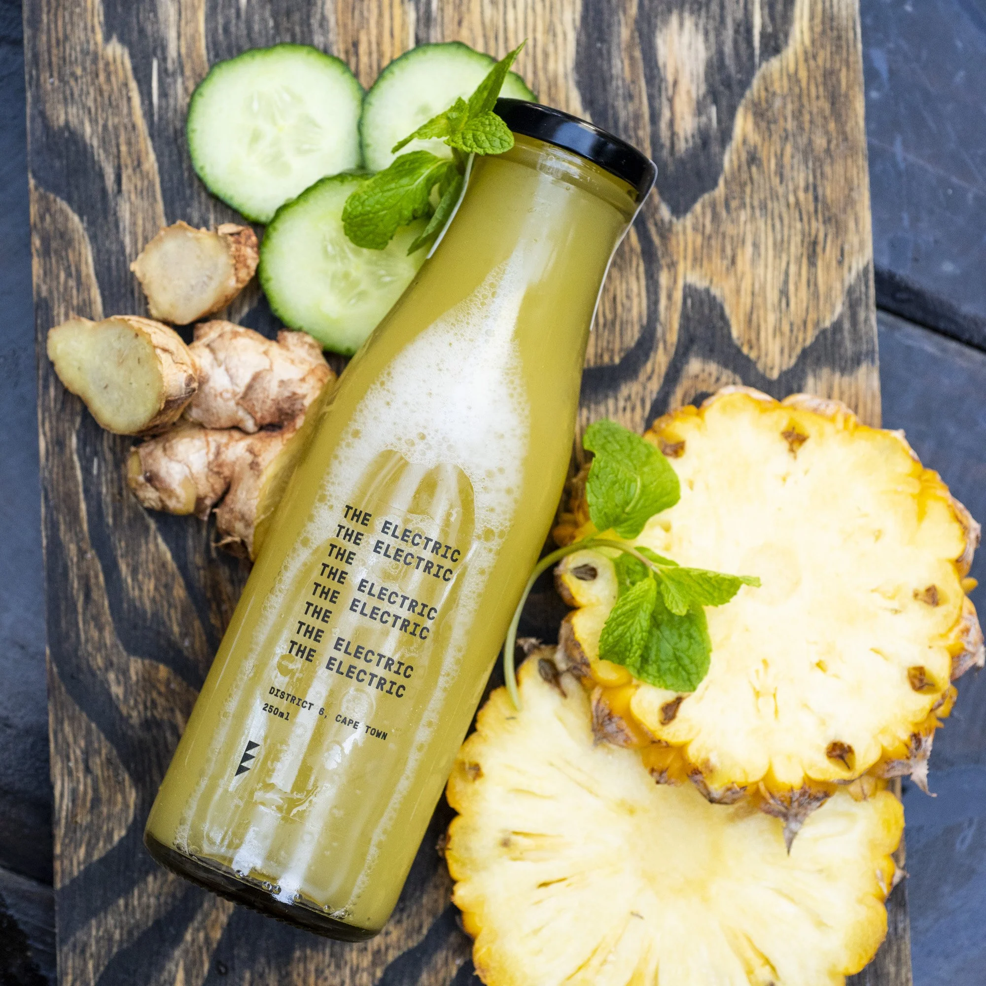 A glass bottle of yellow-green juice labeled 'The Electric' placed on a wooden surface, surrounded by sliced cucumbers, pineapple, ginger, and fresh mint leaves.