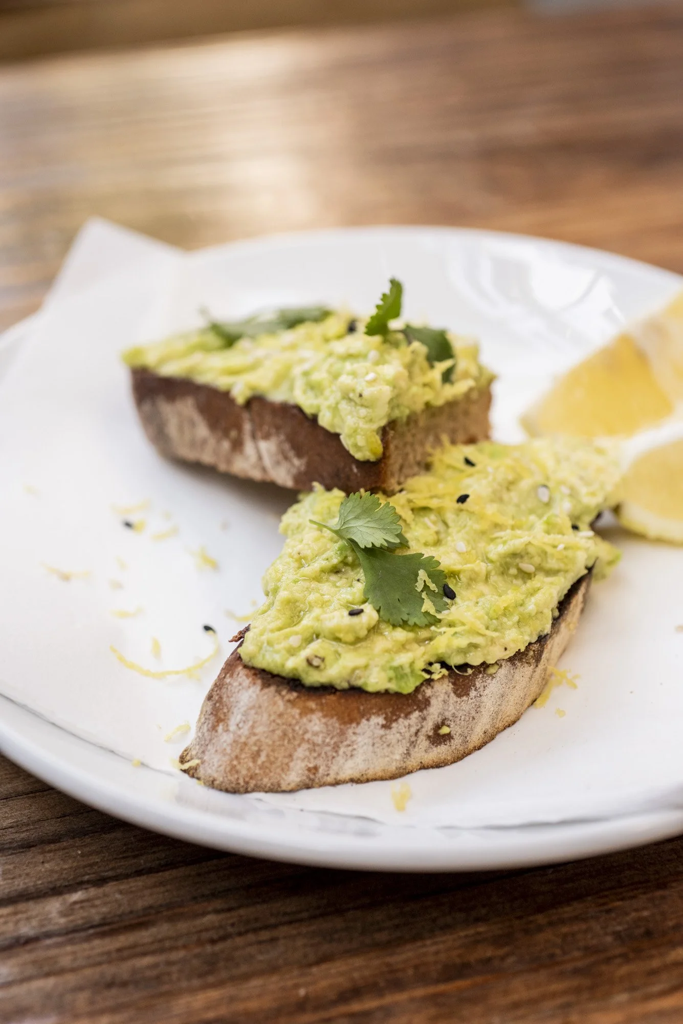 Two slices of toasted bread with mashed avocado, garnished with cilantro, on a white plate with lemon wedges on the side, placed on a wooden table.