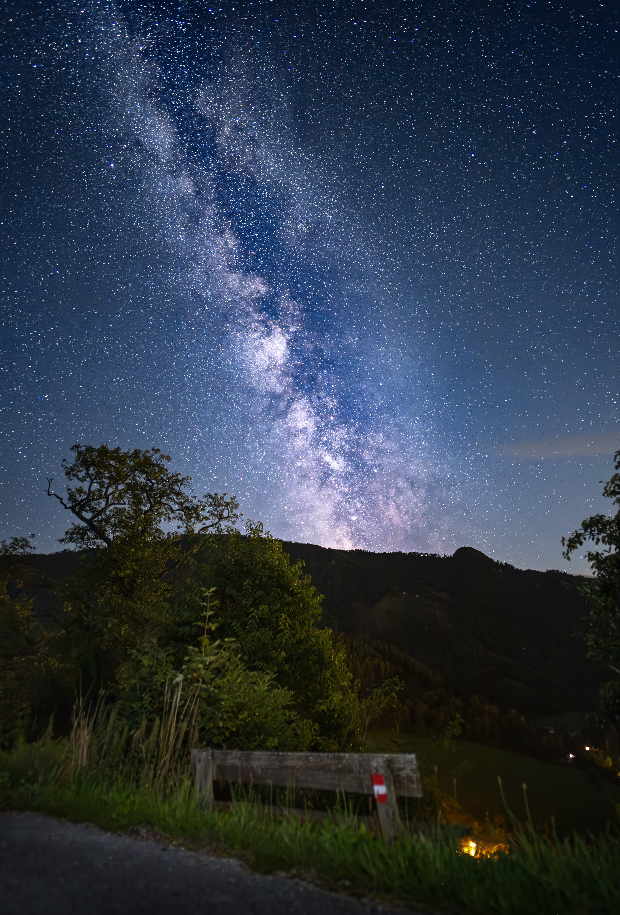 Sternenklare Nacht mit sichtbarster Milchstraße über Hügel und Bäumen, mit Bänken und Wiese im Vordergrund.