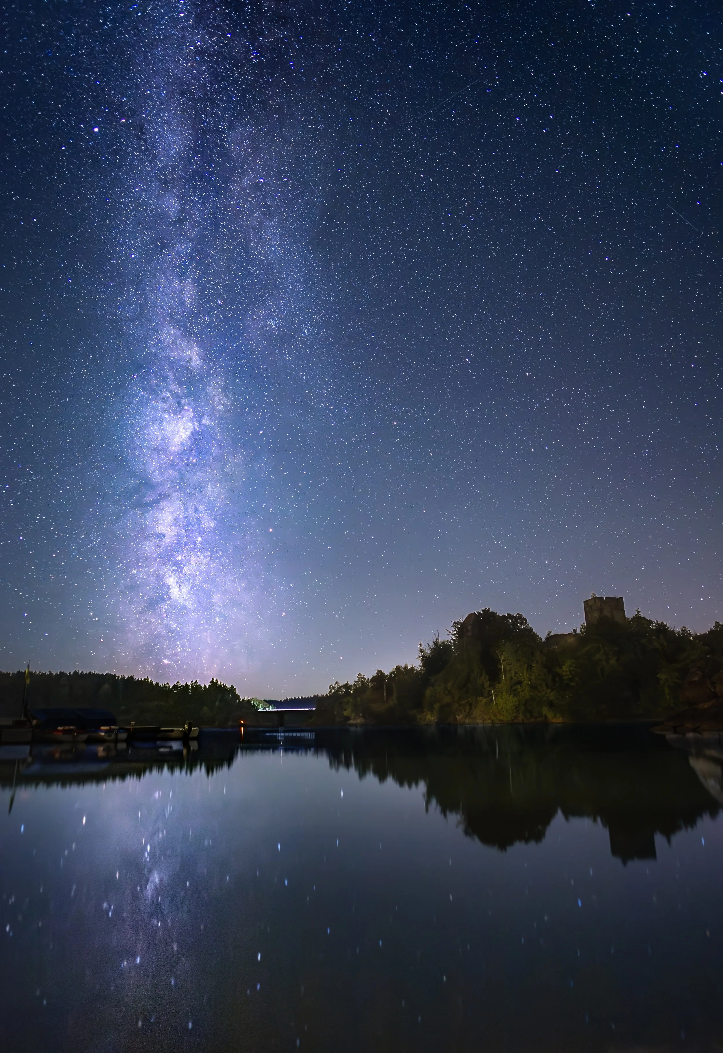 Sternenhimmel und Milchstraße über einem See mit Booten und einem Hügel mit einer Burg an der Küste bei Nacht.