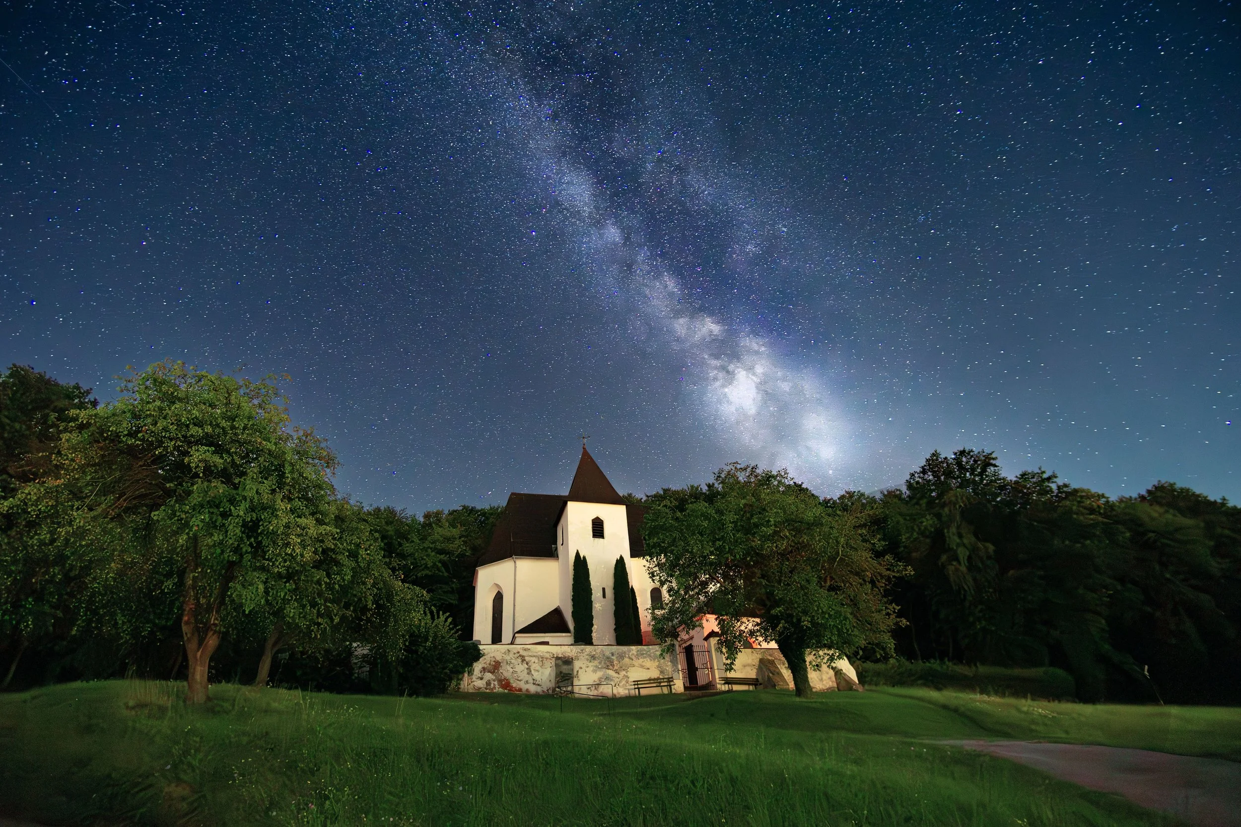 Sternenklare Nacht über einer weißen Kirche inmitten grüner Bäume auf einer Wiese.