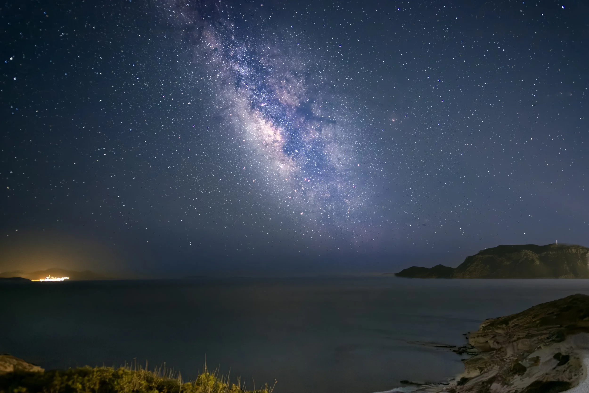 Sternenhimmel mit sichtbarster Milchstraße über dem Meer bei Nacht, umgeben von Hügeln und einer Küstenlinie.