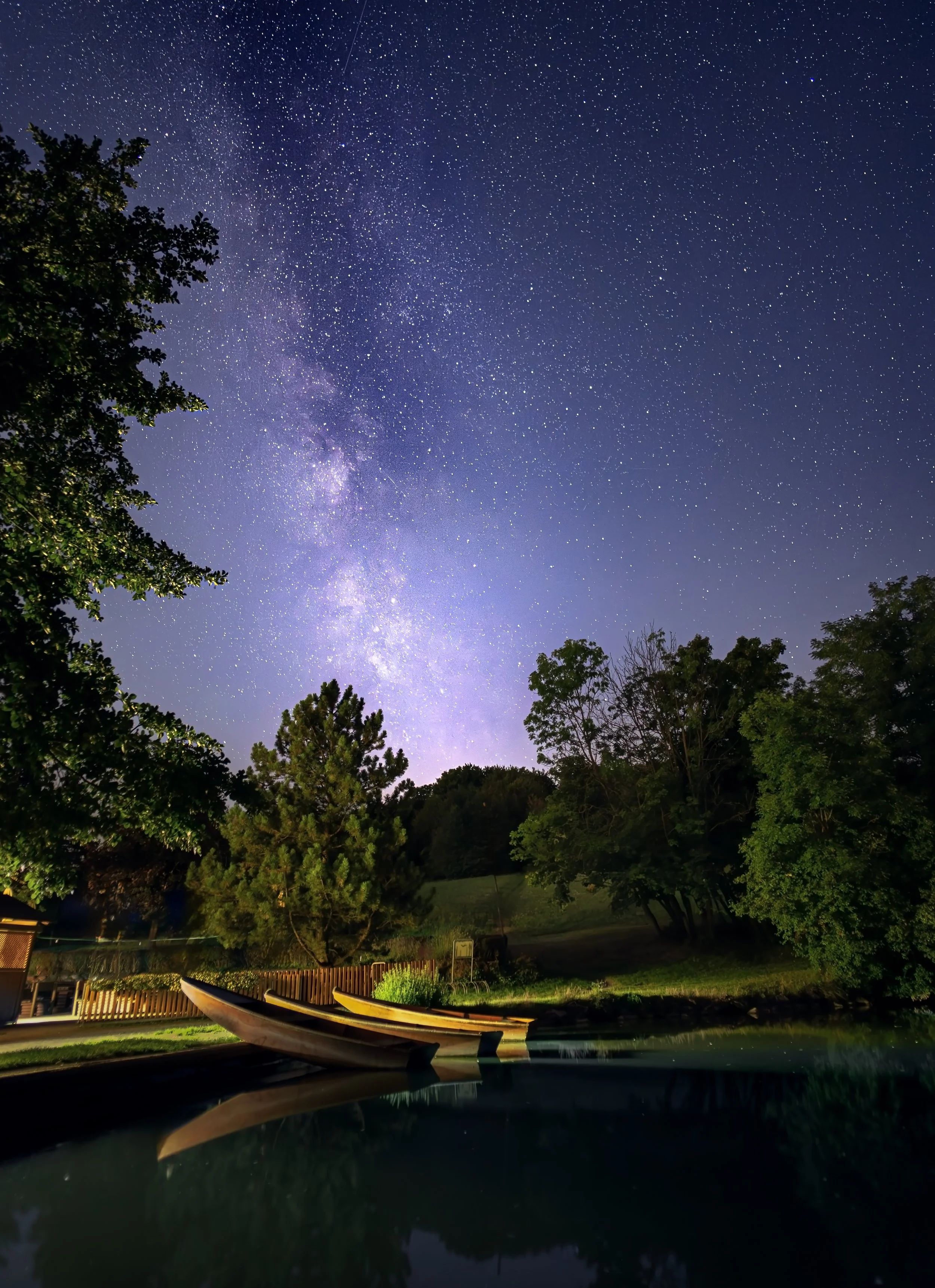 Sternenübersätes Nachthimmel mit Blick auf den Himmel über einem See mit mehreren Booten, Bäumen und einem Zaun.