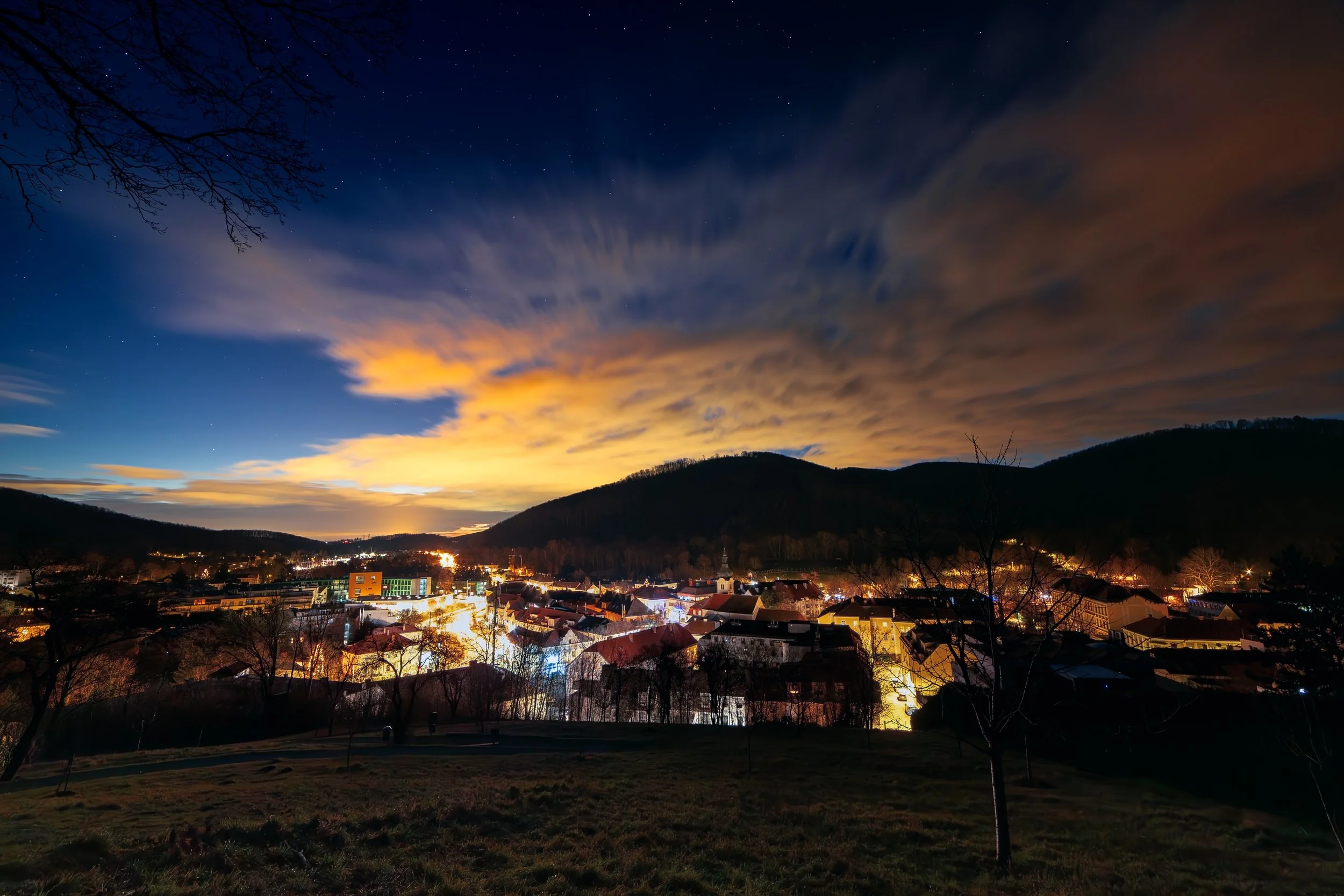 Abendliche Stadtansicht mit beleuchteten Gebäuden, Bergen im Hintergrund, Wolken am Himmel und Bäumen im Vordergrund.