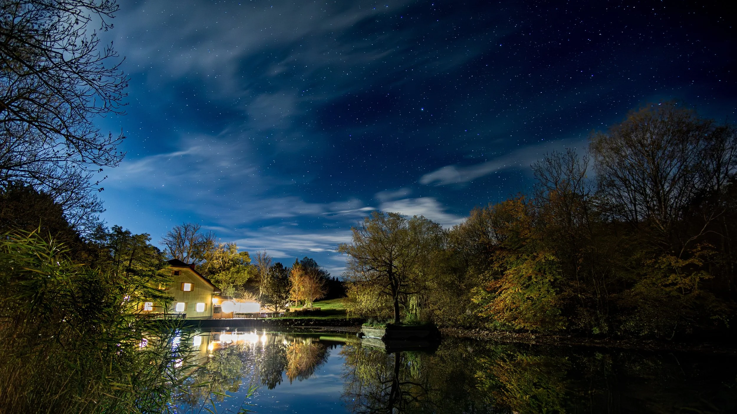 Sternenklare Nacht über einem Wassersee, umgeben von Bäumen, mit einem Haus auf der linken Seite, das von Lampen beleuchtet wird, und Wolken am Himmel.