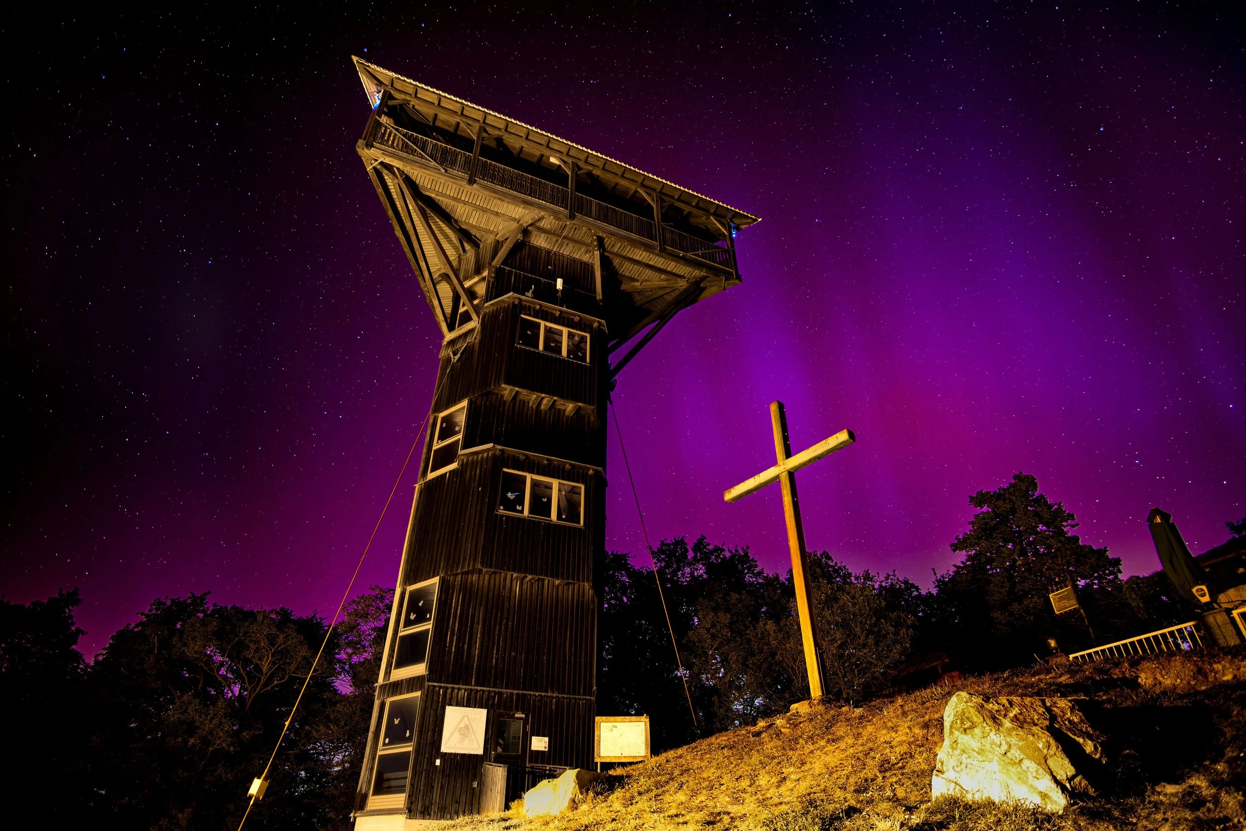 Ein Holz-Wartturm mit mehreren Fenstern und eine große Kreuz neben einem Hügel bei sternenbedecktem Himmel mit purplefarbener Aurora.