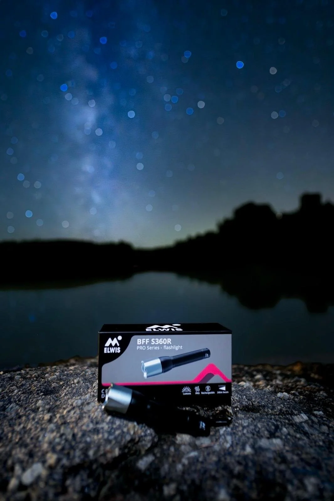 Eine Taschenlampe, ELWIS BFF S360R PRO Series, liegt auf einem felsigen Boden vor einer Verpackung in der Nähe eines Sees bei Nacht, mit einem Sternenhimmel im Hintergrund.