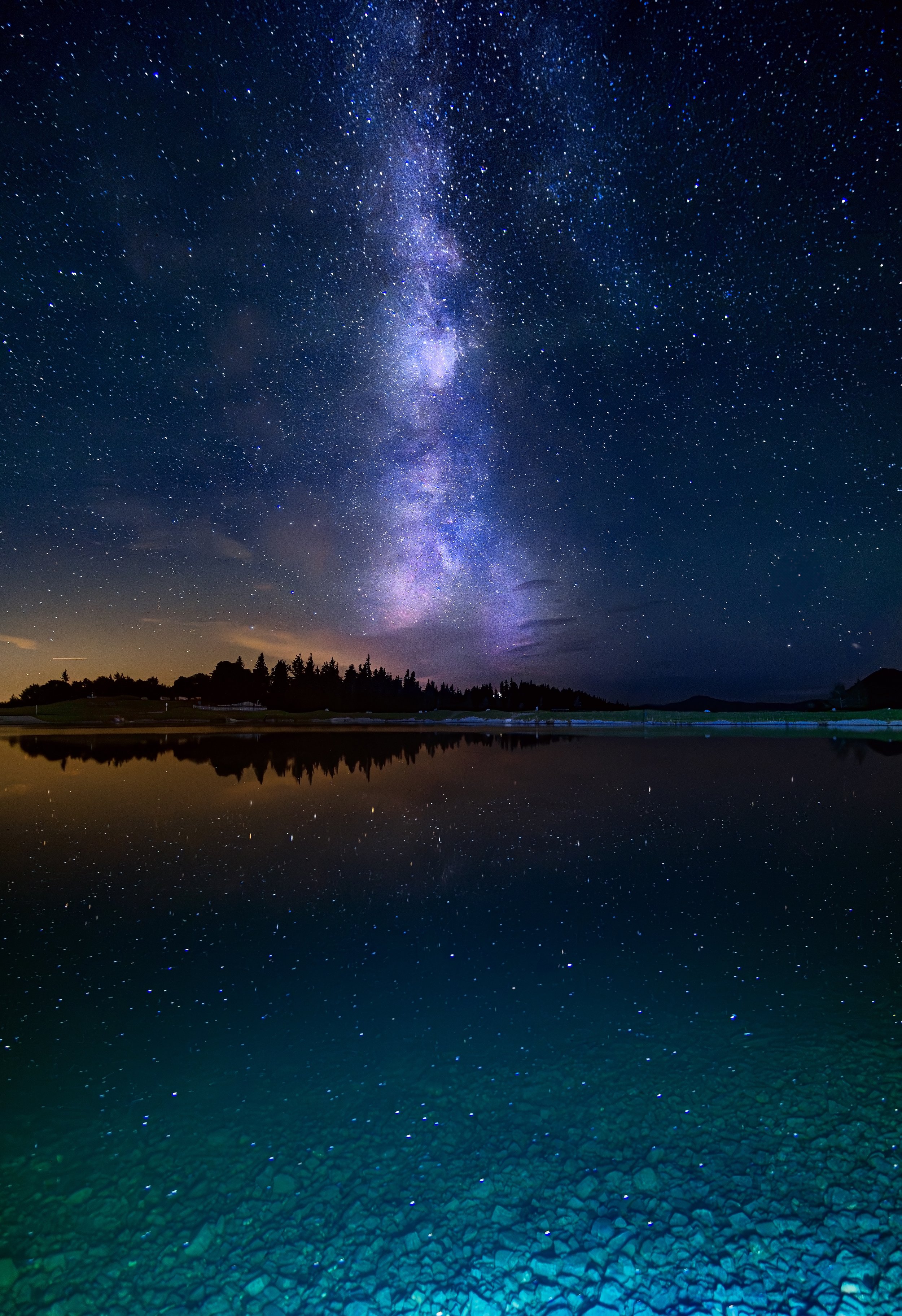 Sternenklare Nacht über einem See mit spiegelnder Wasseroberfläche, galaktischer Himmel und Silhouetten von Bäumen am Horizont.