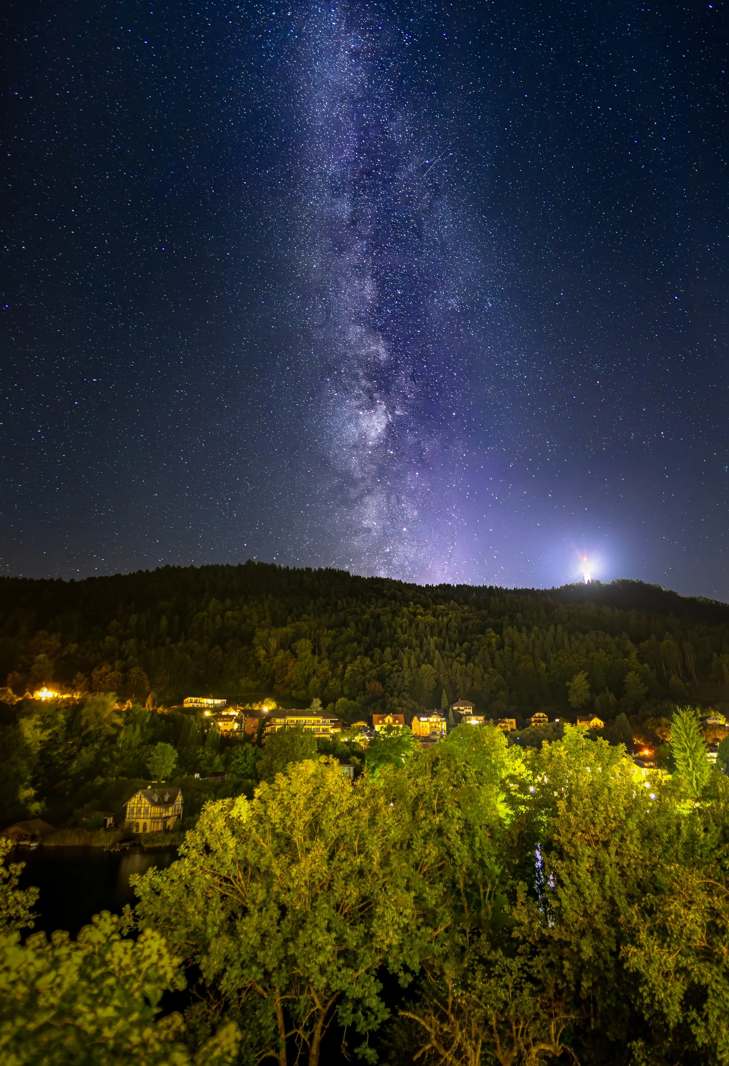 Sternenklare Nacht über einem Dorf mit Hügel, Bäumen und einer Lichterkette.