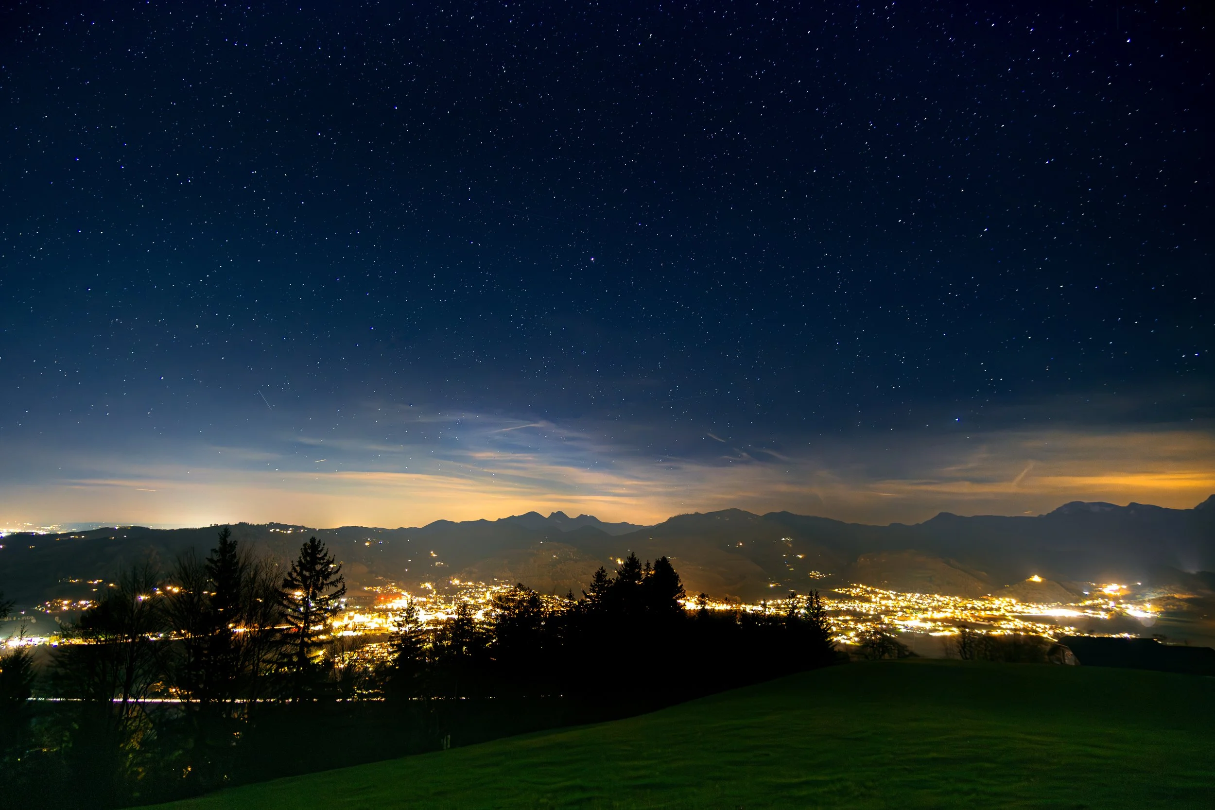 Sternenhimmel über einer Stadt in den Bergen bei Nacht, mit sichtbaren Sternkonstellationen und leuchtender Stadtbeleuchtung im Tal.