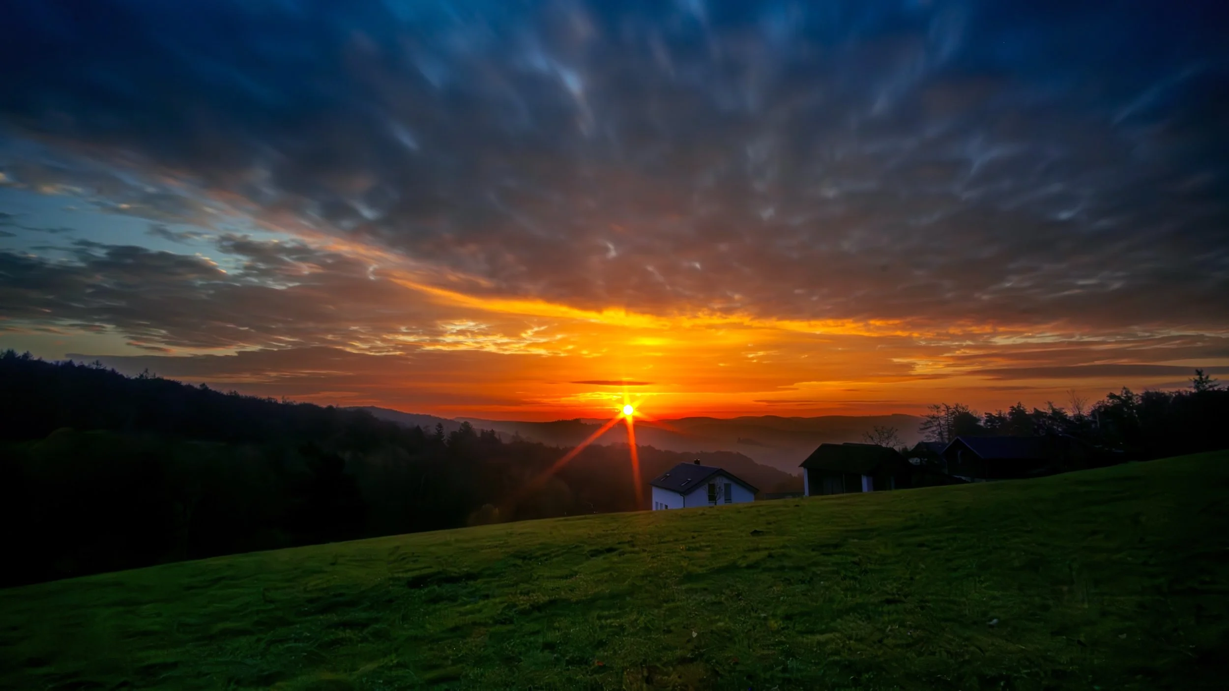 Ein Sonnenuntergang über einer grünen Wiese mit einigen Häusern im Vordergrund und bewölktem Himmel
