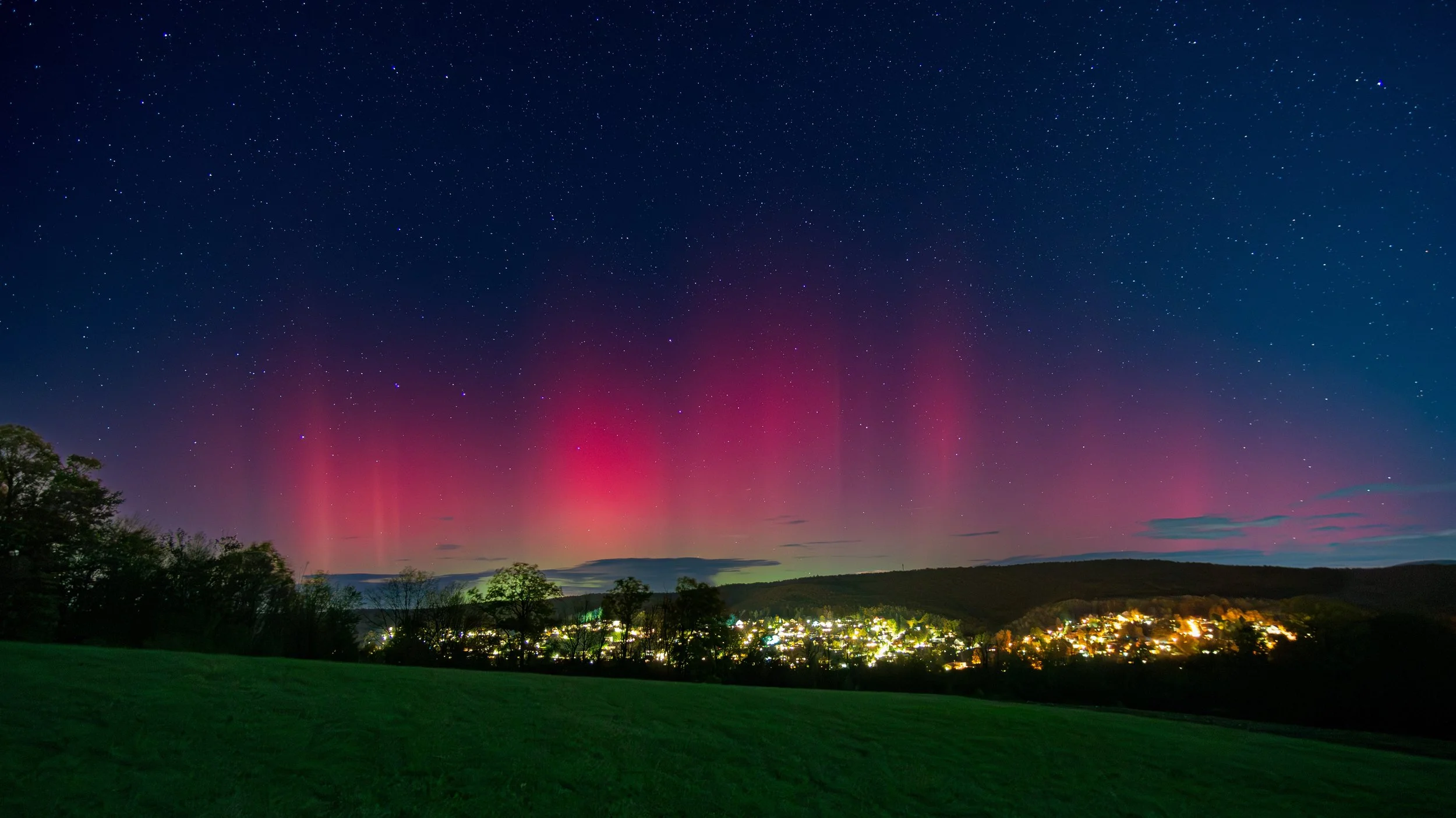 Nordlichter über einer kleinen Stadt bei Nacht, mit grüner Wiese im Vordergrund und einem Sternenhimmel.
