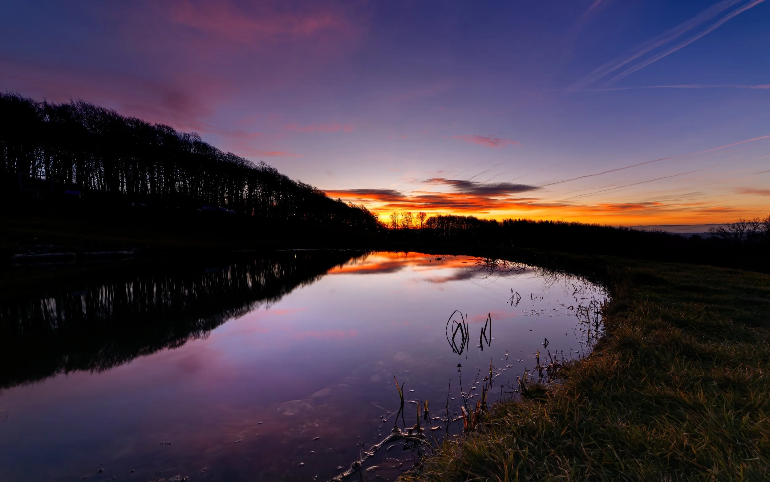 Ein malerischer Sonnenuntergang über einem ruhigen Fluss, mit reflektierendem Himmel in pink, orange und blau, umgeben von Bäumen und Gras, bei Dämmerung.
