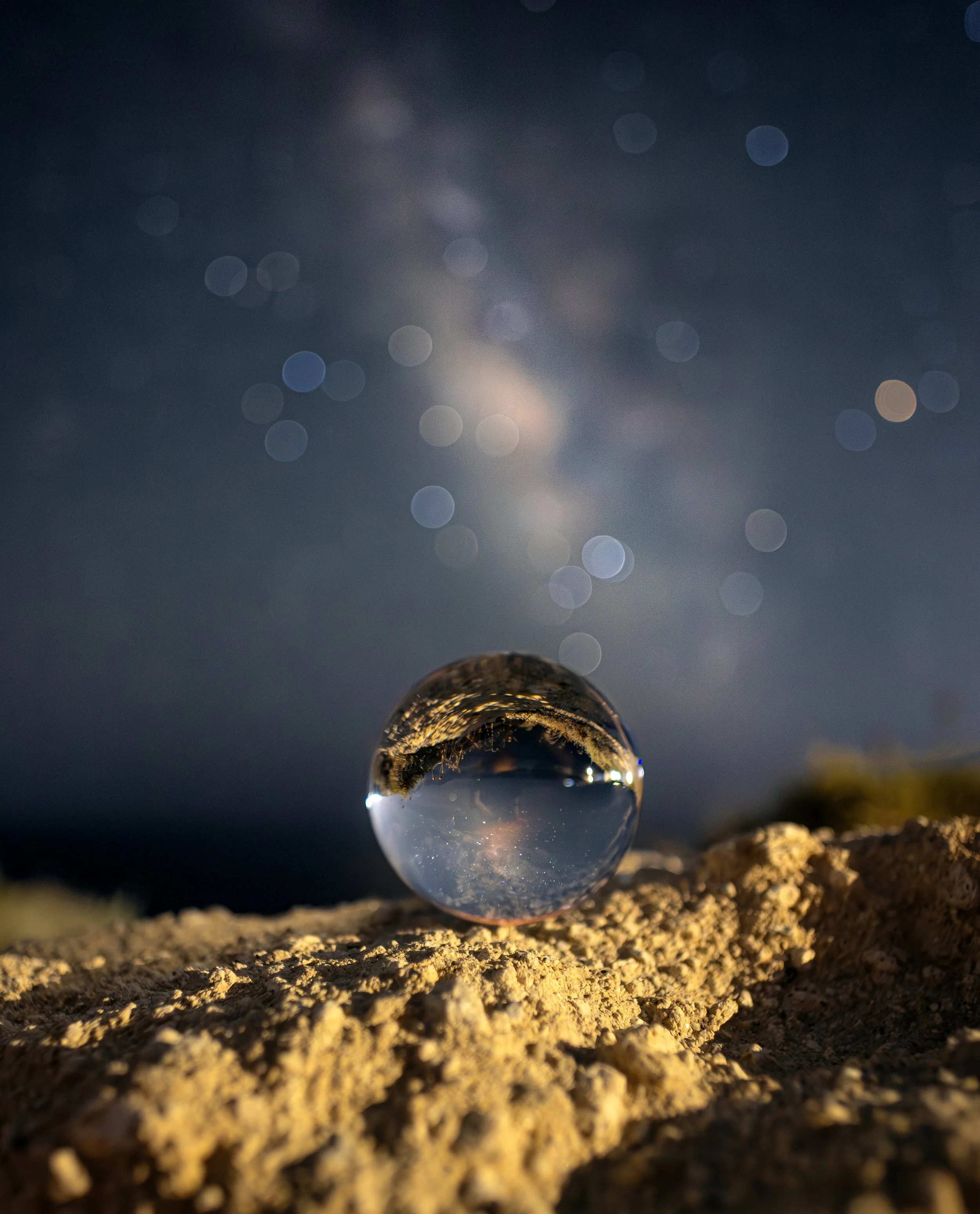 Hier ist ein Glasglobus auf sandigem Boden, das die Umgebung, den Himmel und die Sterne reflektiert, bei Nacht mit Sternenhimmel im Hintergrund.