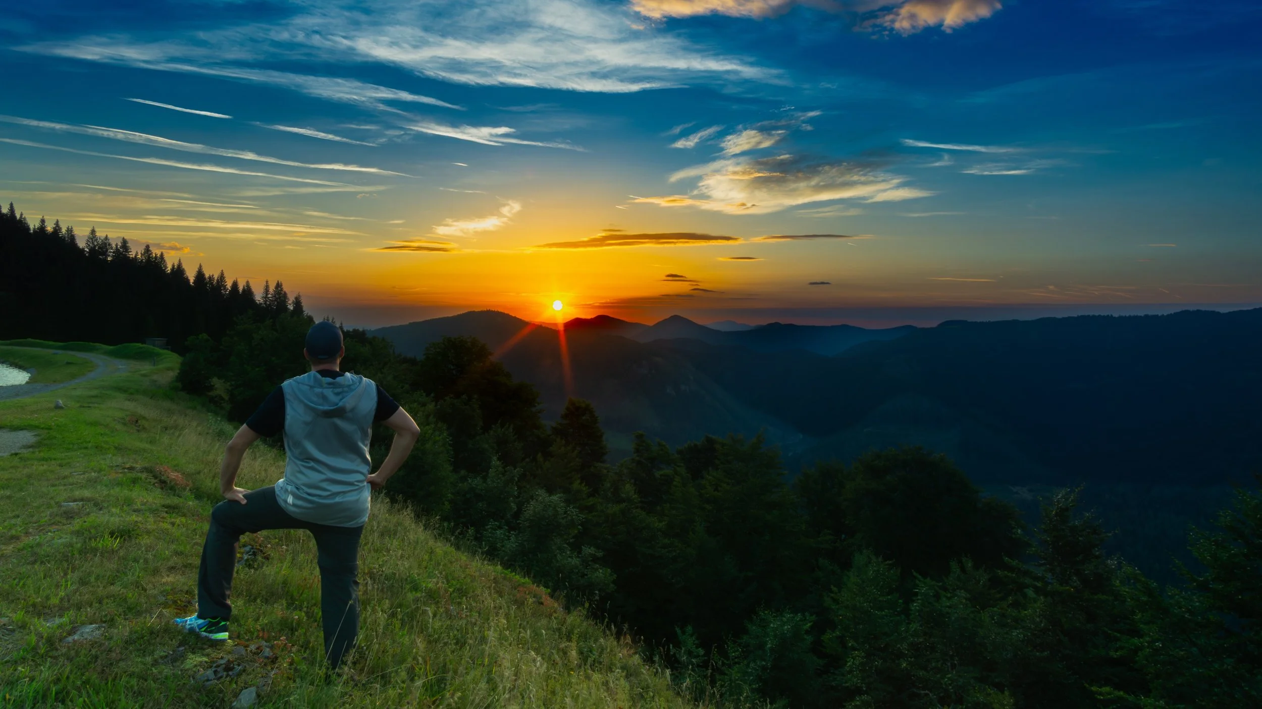 Ein Mann steht auf einer grünen Wiese und schaut auf einen Sonnenuntergang über den Bergen, umgeben von bewaldeten Hügeln.