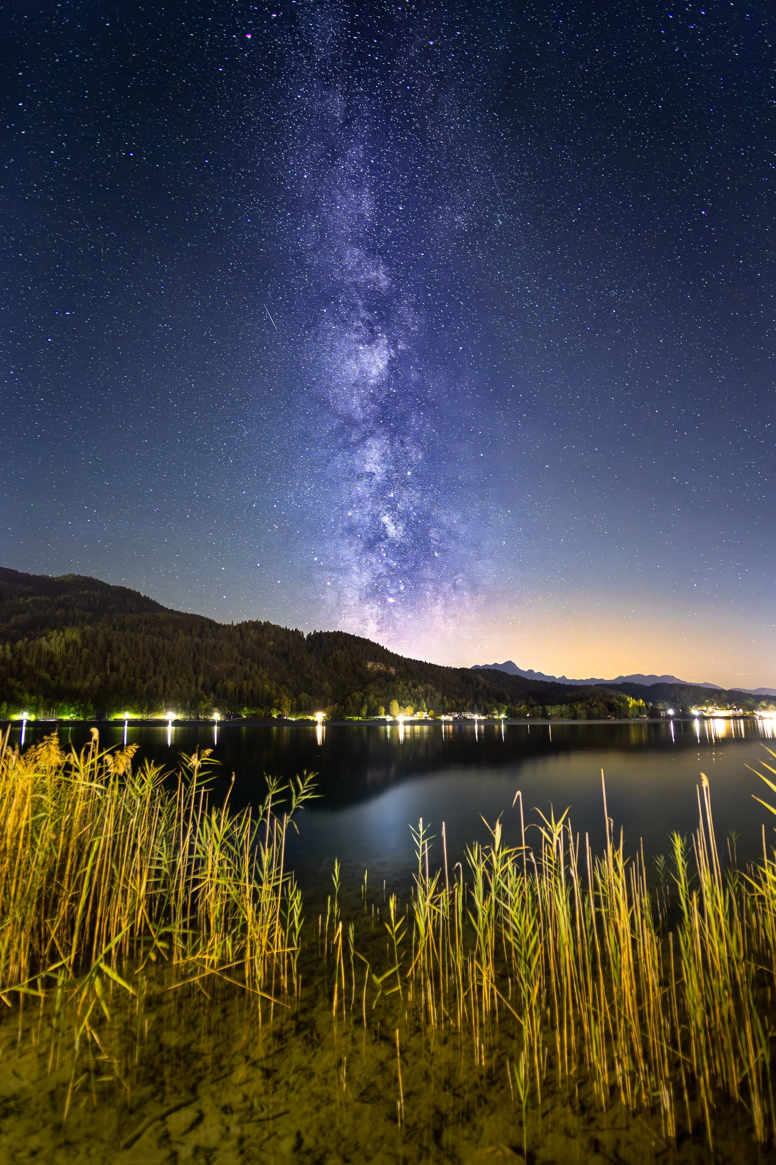 Sternenklare Nacht über einem ruhigen See mit Bergen im Hintergrund und Gräsern im Vordergrund.