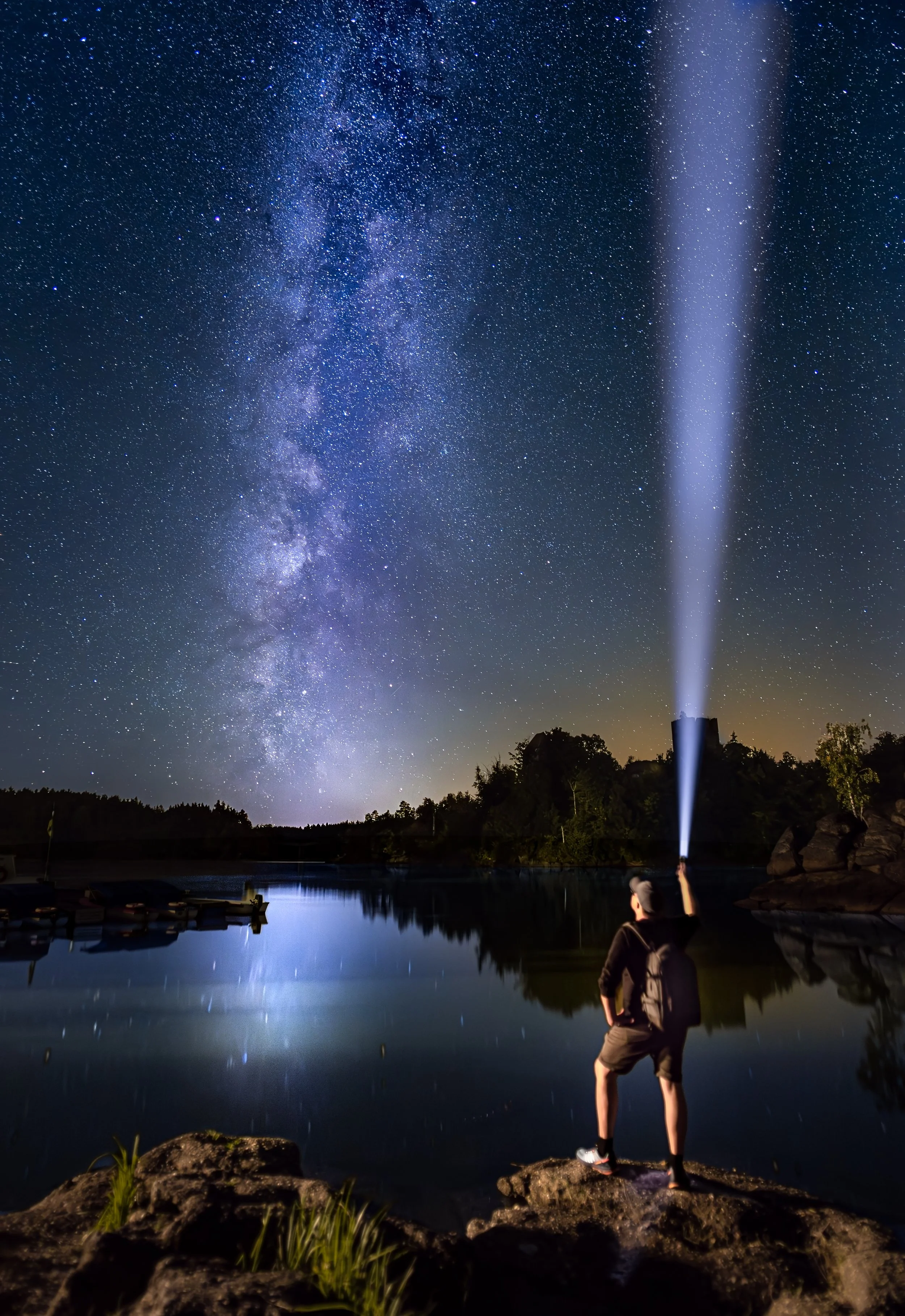 Ein Mensch steht am Ufer eines Sees in der Nacht und leuchtet mit einer Taschenlampe in den Himmel, der voller Sterne und der Milchstraße ist.