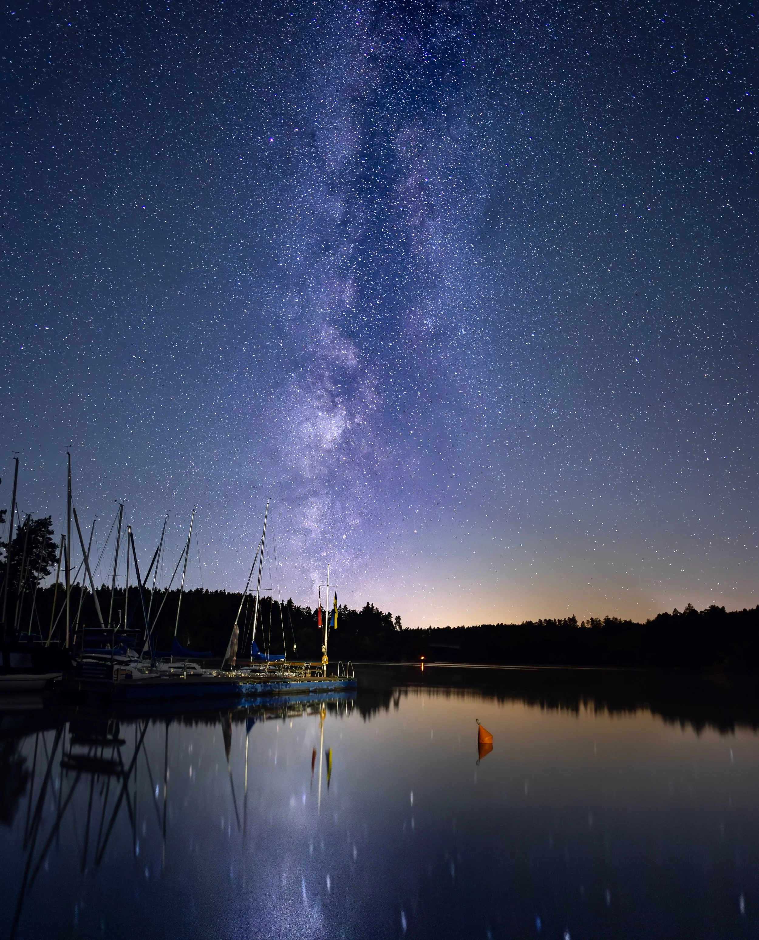 Sterne leuchtender Nachthimmel über einem ruhigen See mit Booten, Bäumen am Horizont und hellen Reflexionen im Wasser.