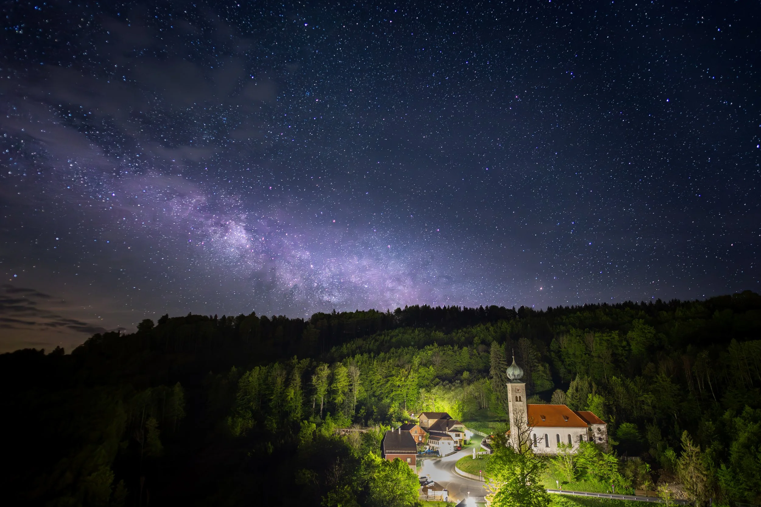 Sternenhimmel über einer kleinen, beleuchteten Dorfkirche mit rotem Dach und umgebenen Häusern, umgeben von bewaldeten Hügeln.