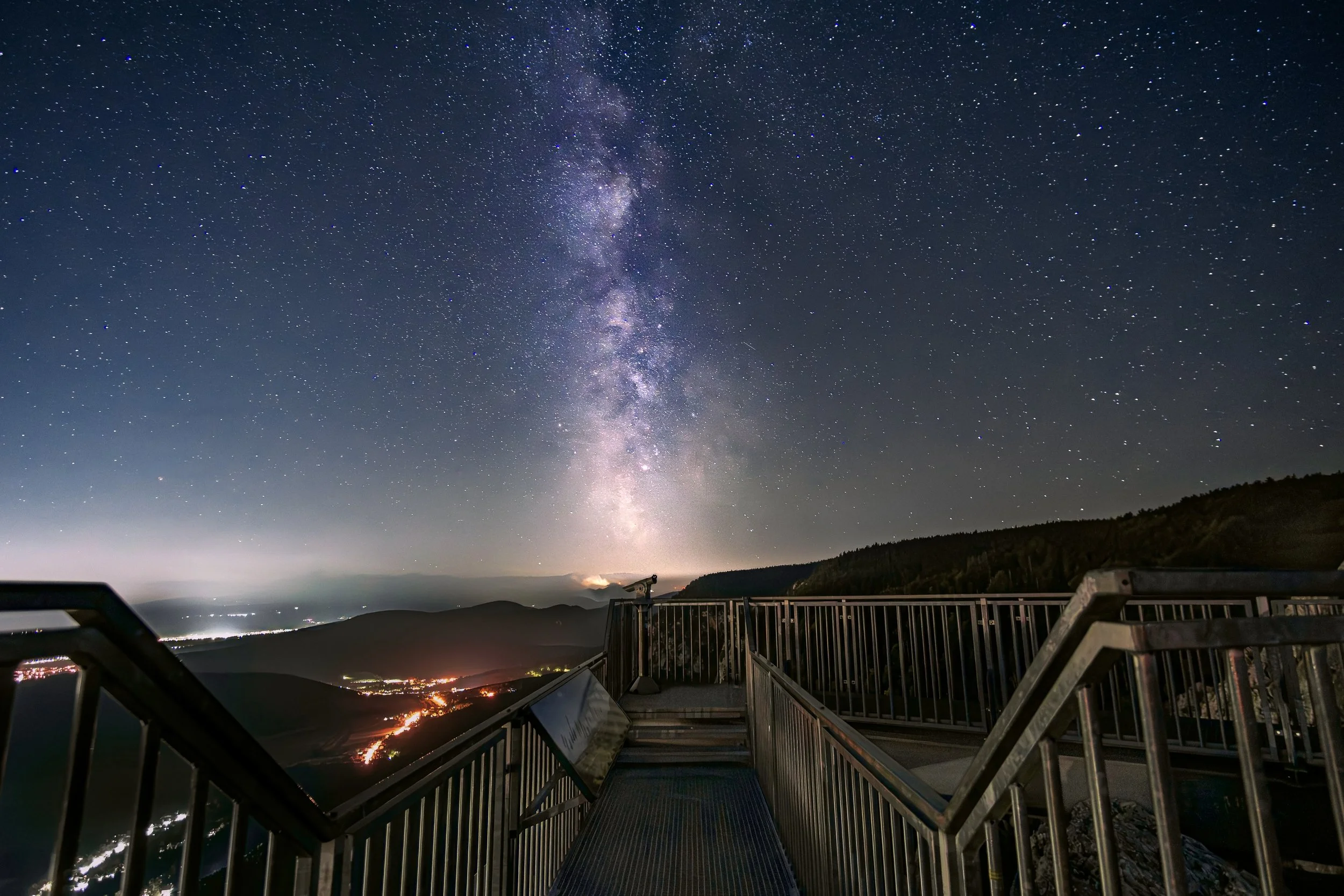 Sternenklare Nacht am Aussichtspunkt mit Metalldielen und Geländer, Blick auf den Himmel mit Milchstraße, Hügel und Städte im Hintergrund.