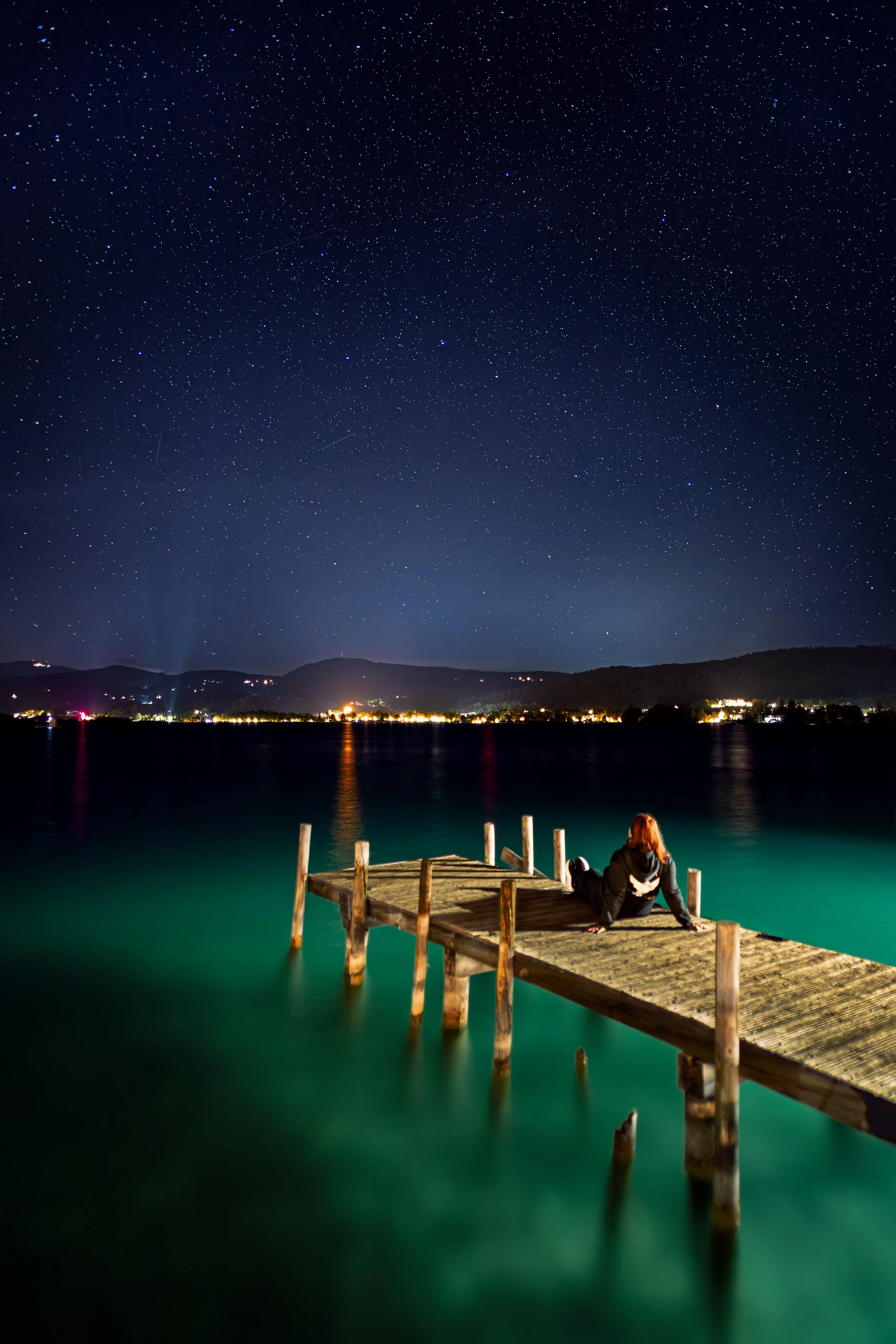 Eine Frau sitzt auf einem Steg am Wasser und blickt in den Sternenhimmel bei Nacht, mit glitzernden Sternen über einem ruhigen See. Im Hintergrund sind die Lichter einer Stadt sichtbar.