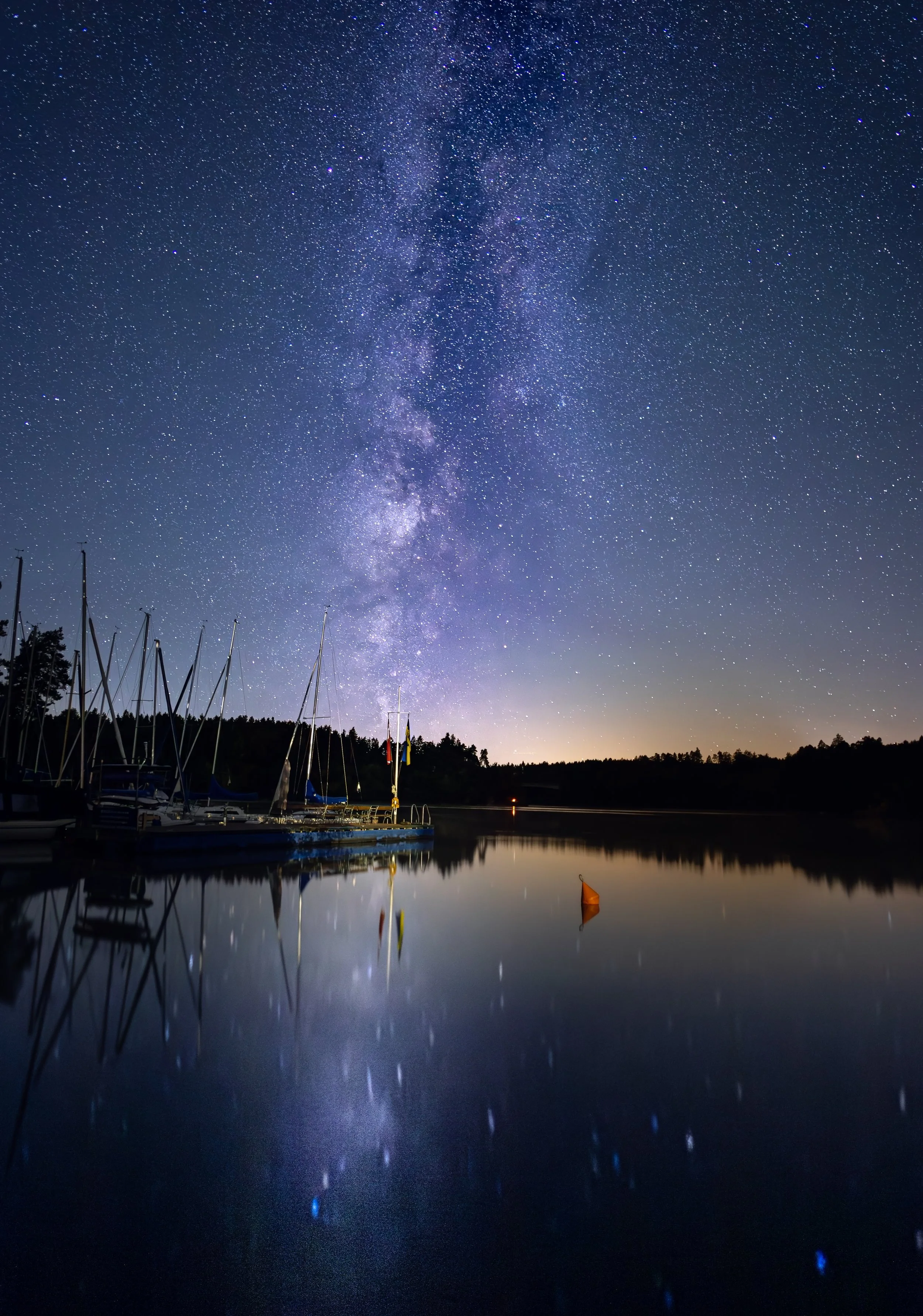 Sterne und die Milchstraße spiegeln sich im ruhigen Wasser eines Sees mit Segelbooten am Ufer bei Nacht.