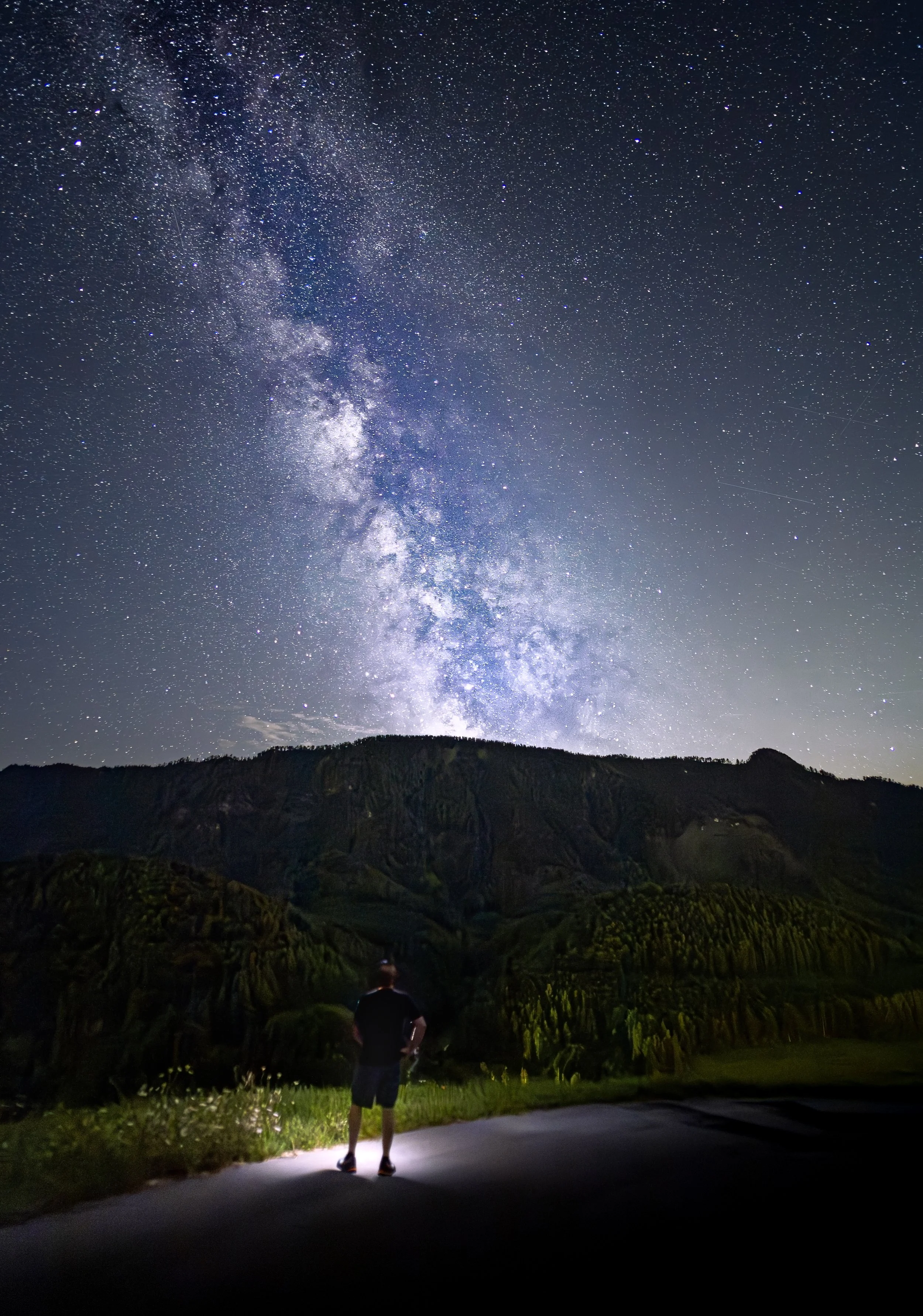 Ein Mensch steht nachts auf einer Straße und schaut in den Sternenhimmel mit der Milchstraße über hügeligen Landschaften.