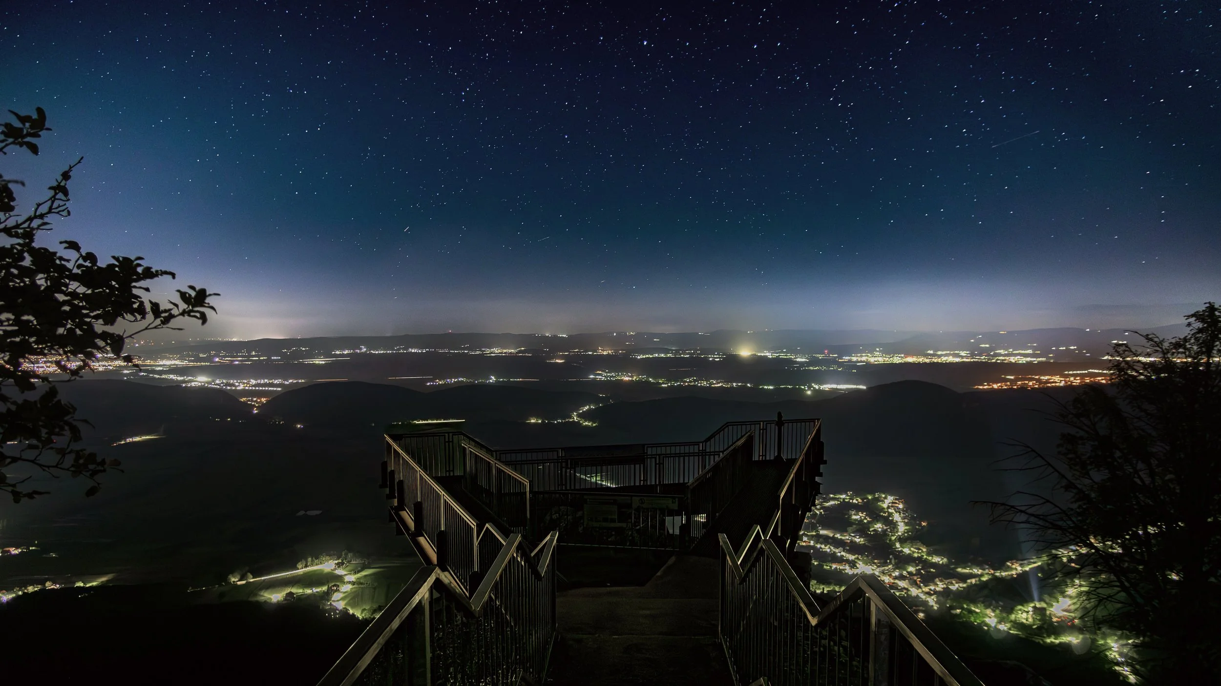 A starry night sky über einer Berglandschaft mit einer Aussichtsplattform aus Holz und Metall im Vordergrund, die nach unten in eine bewaldete Gegend und mehrere beleuchtete Siedlungen führt.