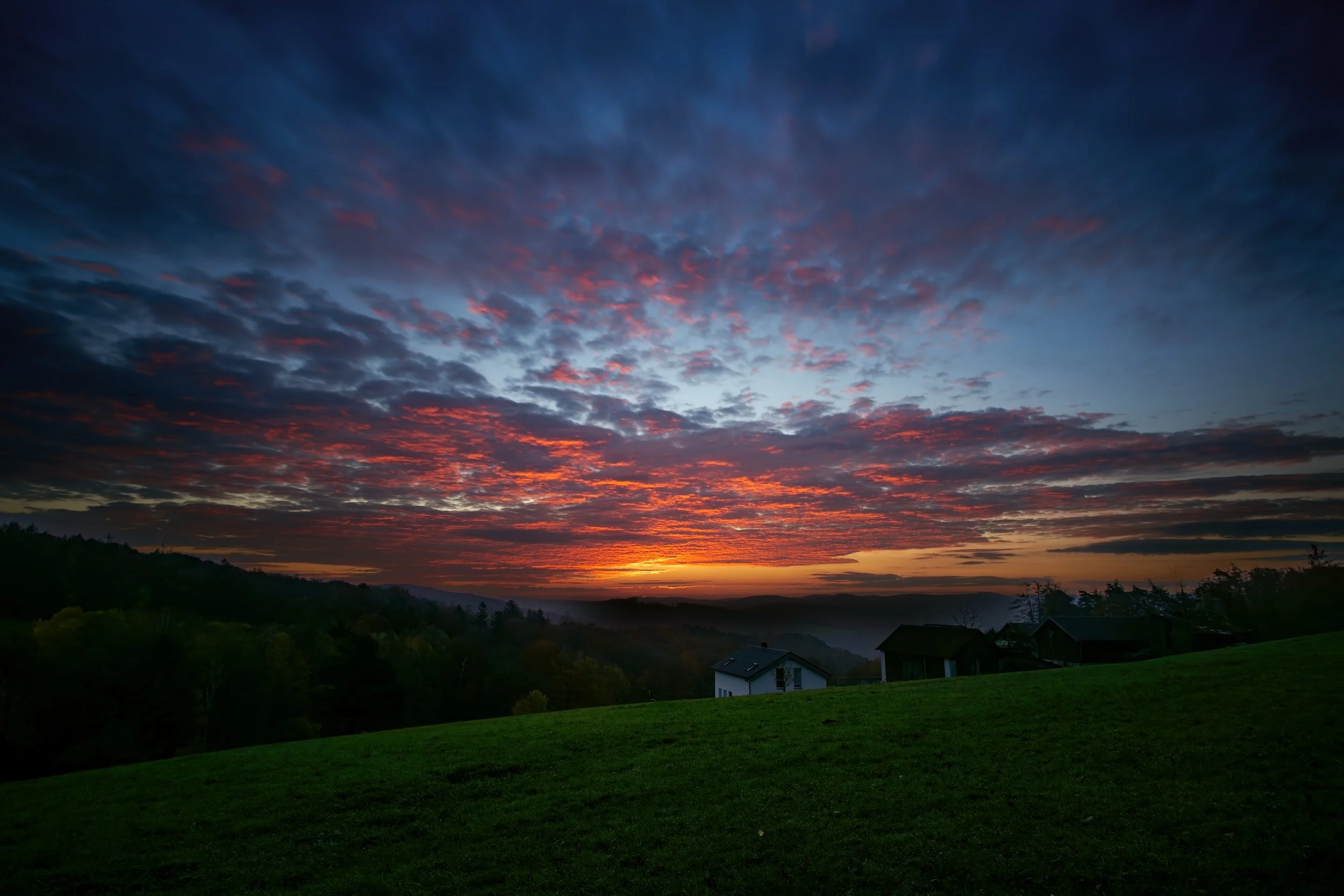 Sonnenuntergang über einer grünen Wiese mit wenigen Häusern, bewölkter Himmel in Rot, Blau und Grau.