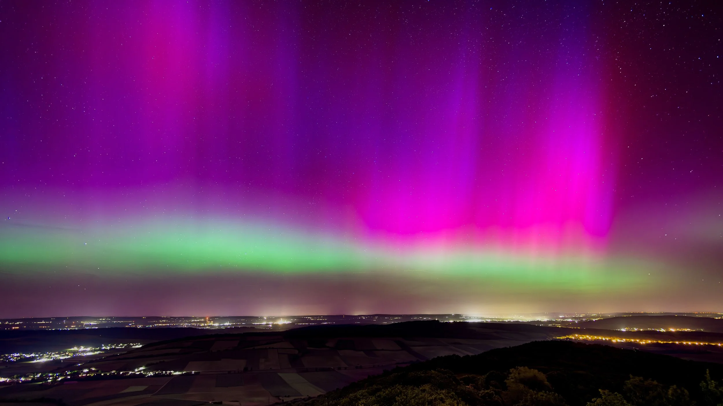 Nordlichter am Nachthimmel in lila, pink und grün, über einer ländlichen Landschaft mit Feldern und leuchtenden Dörfern.