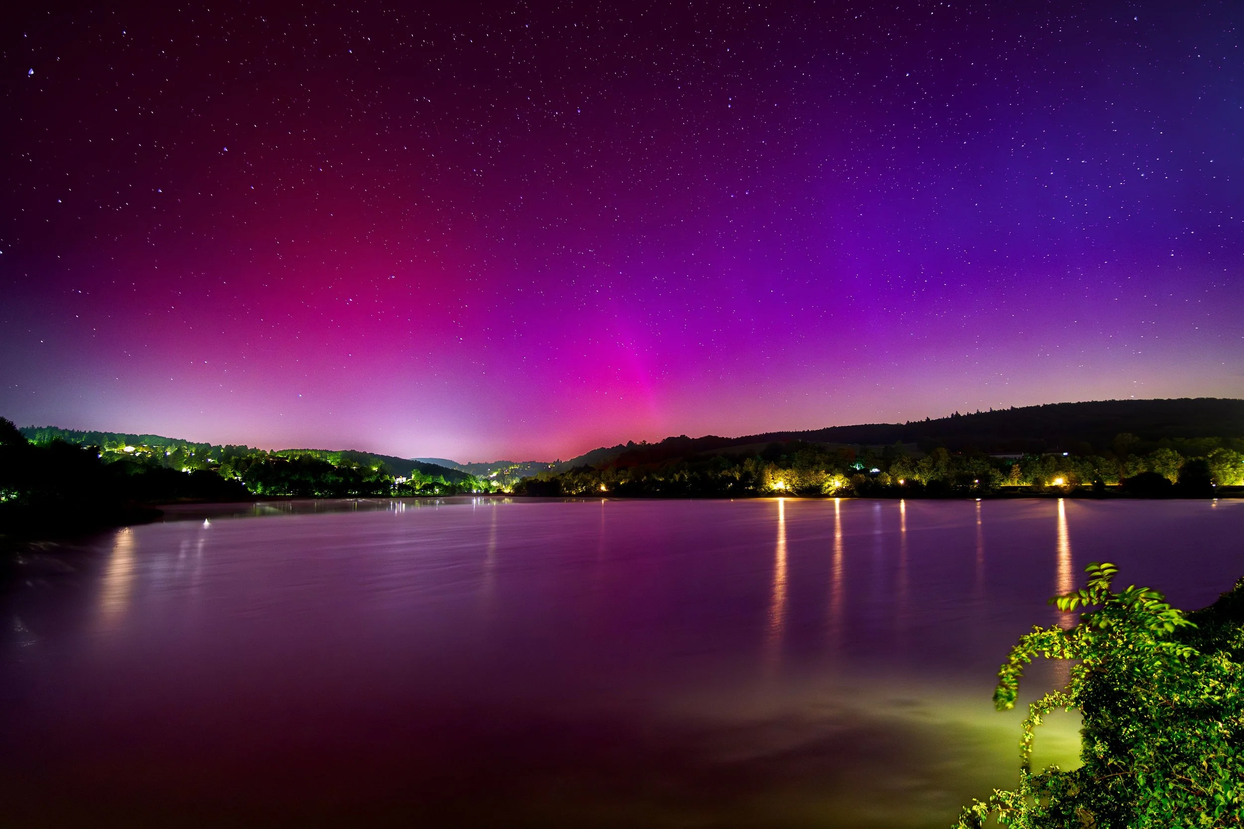 Nachtansicht eines Flusses mit buntem Nordlicht am Himmel, umgeben von bewaldeten Hügeln, reflektiert im Wasser, mit verstreuten Lichtern am Ufer.