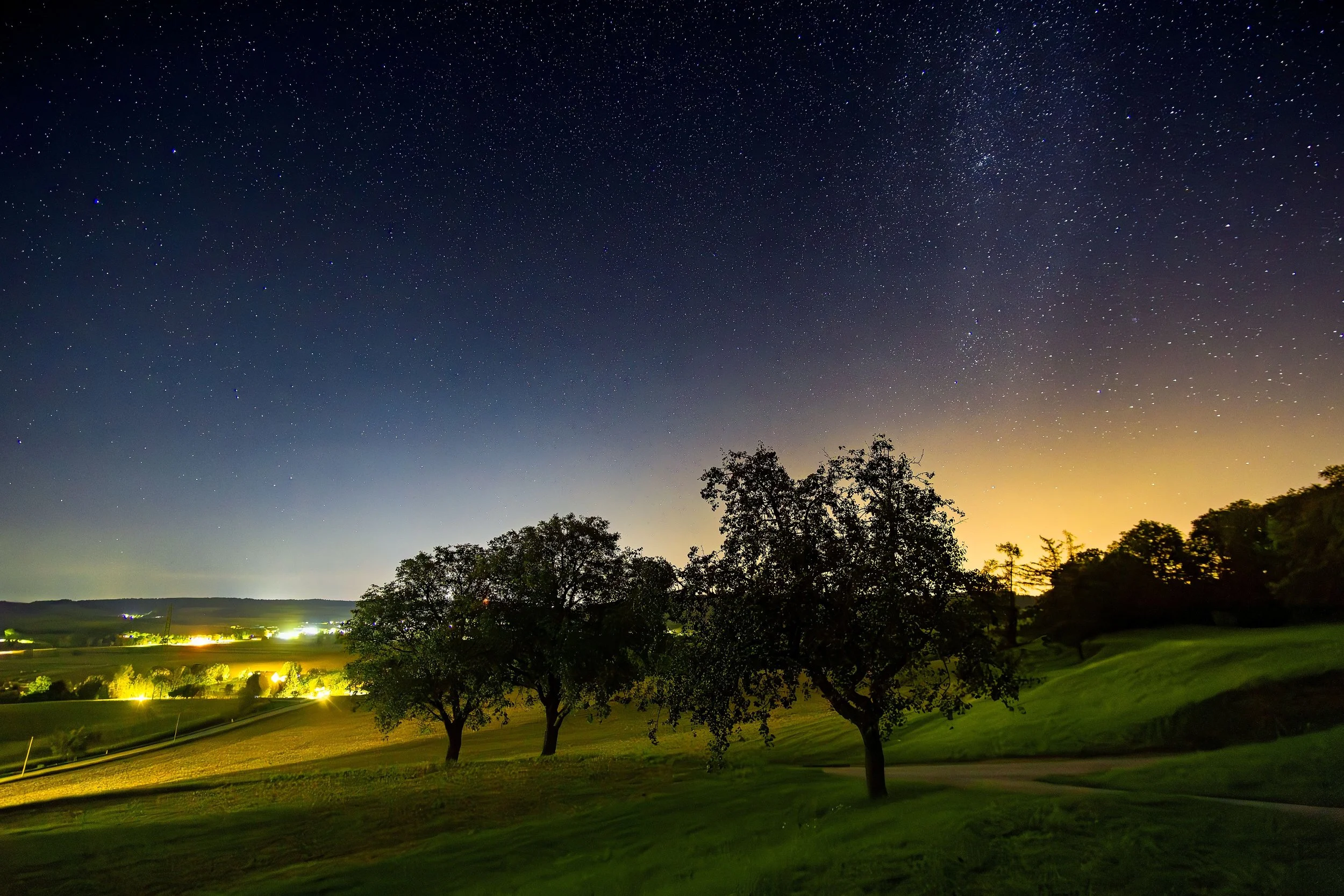 Sternenübersäter Himmel über einem ländlichen Feld mit Bäumen bei Nacht, mit sanfter Beleuchtung im Hintergrund.