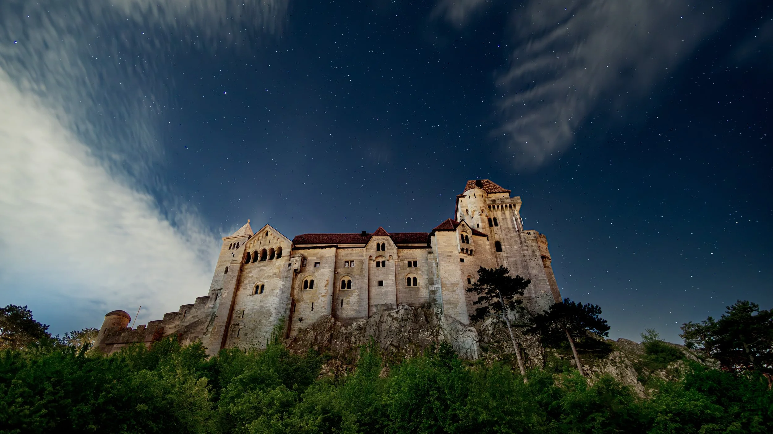 Mittelalterliche Burg auf einem Hügel bei Nacht, umgeben von Bäumen, Sternenhimmel mit Wolken im Hintergrund.