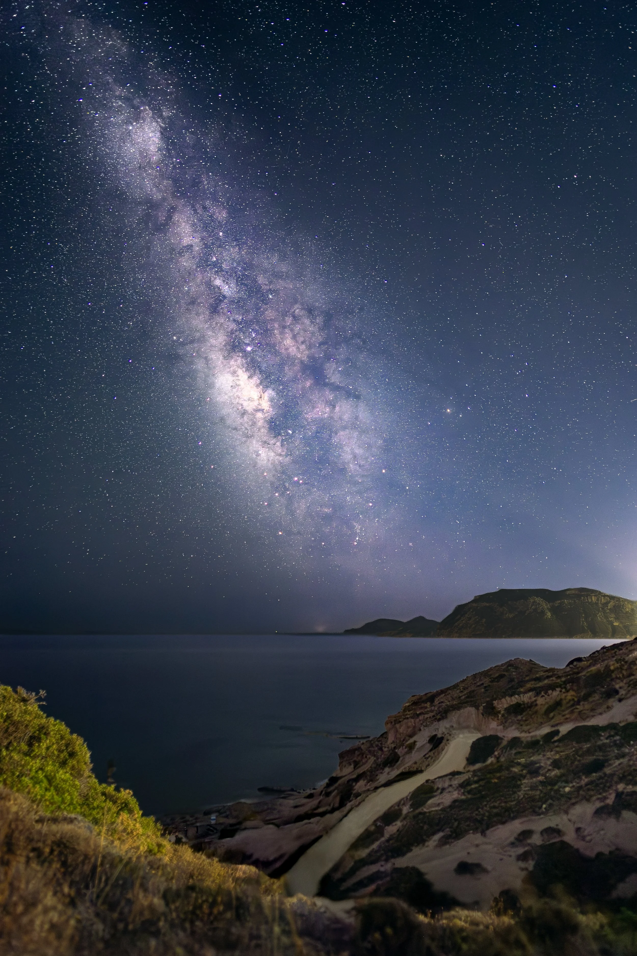 Sternenklare Nacht über einer Küstenlandschaft mit Bergen, Wasser und Felsen im Vordergrund