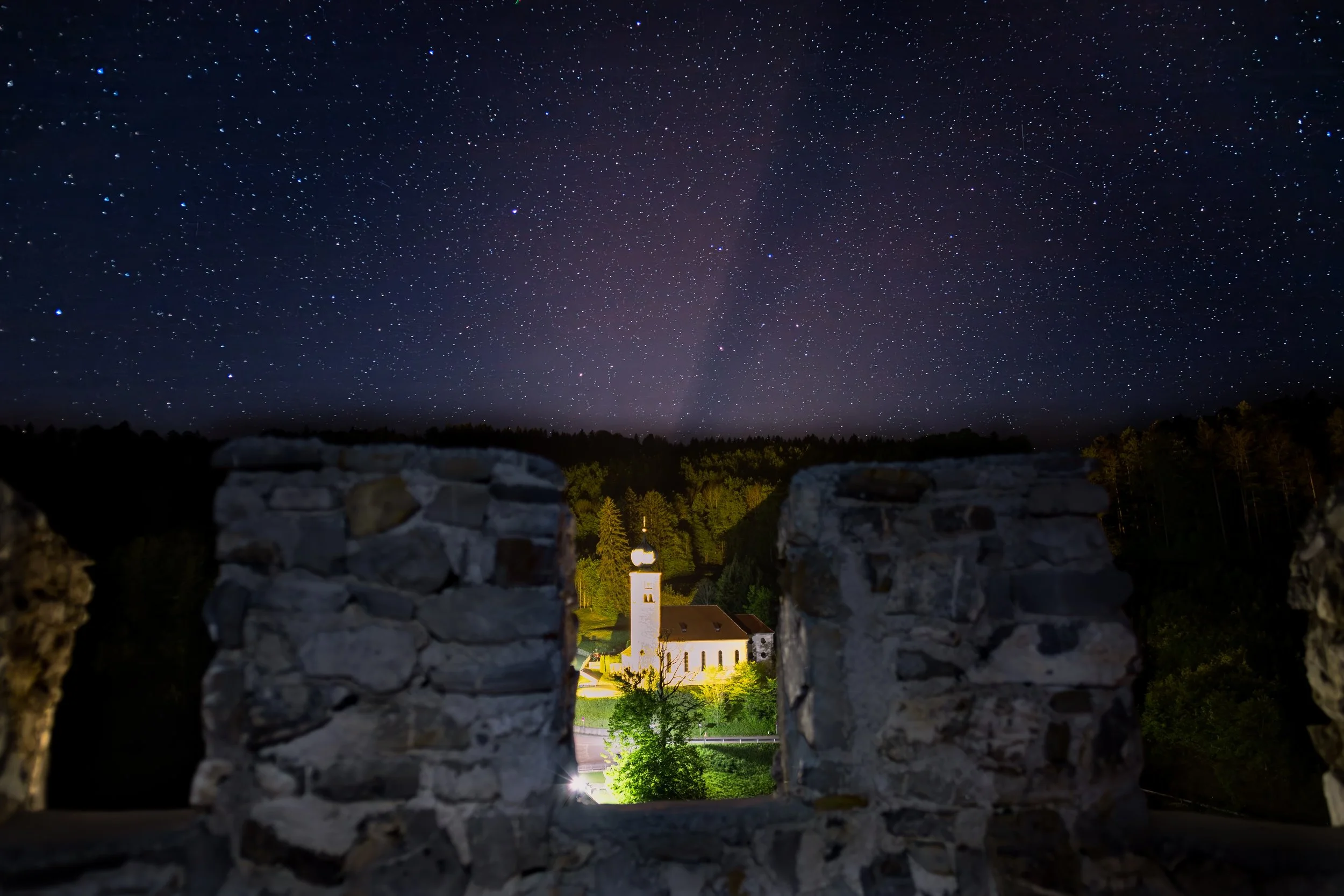 Nachtaufnahme einer kleinen Kirche mit Kirchturm, umgeben von Bäumen, gesehen durch eine alte Steinschleuderöffnung, mit einem sternklaren Himmel im Hintergrund.