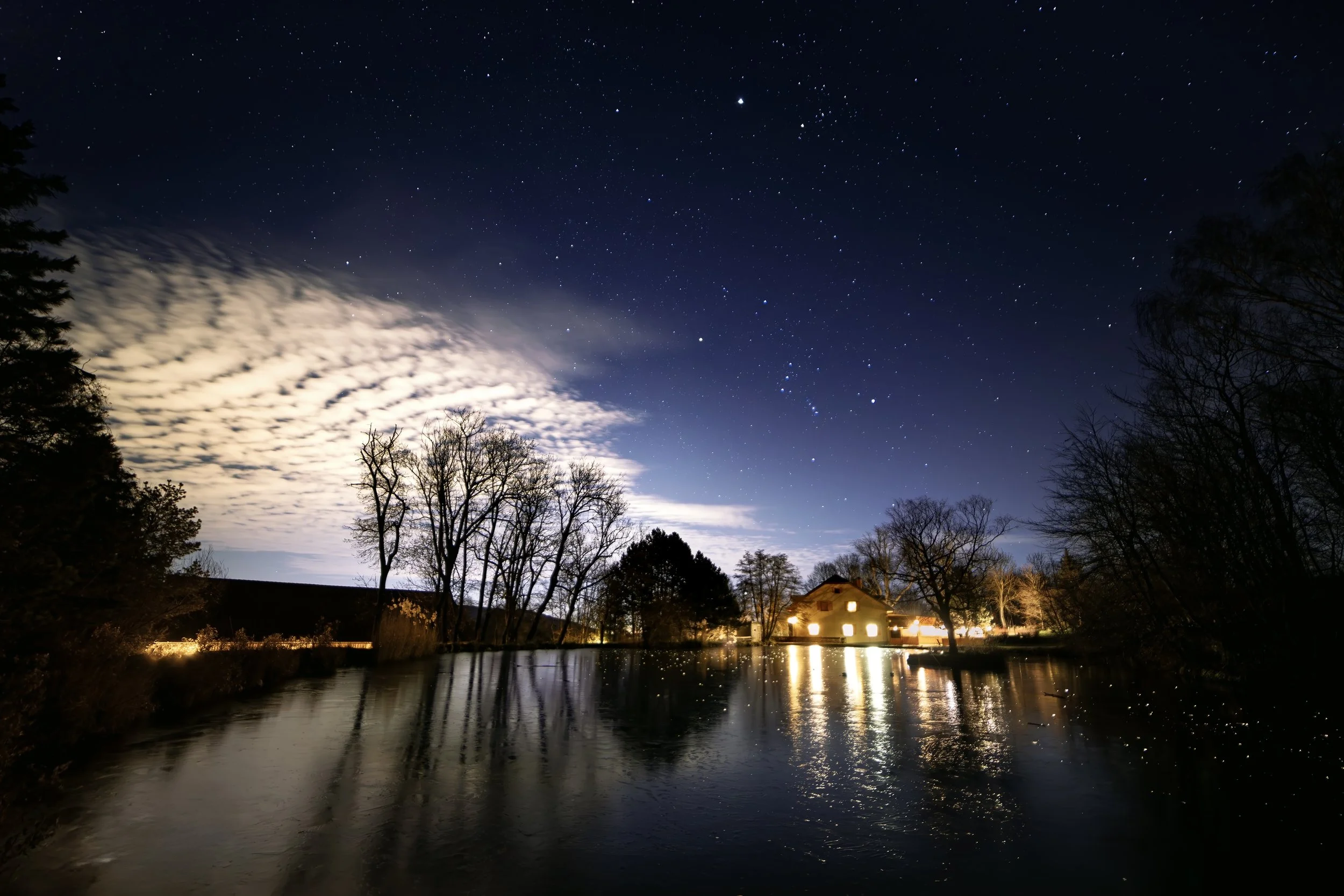 Nachthimmel mit Sternen, Wolken und reflektierendem Wasser, umgeben von Bäumen und einem Haus mit leuchtenden Fenstern