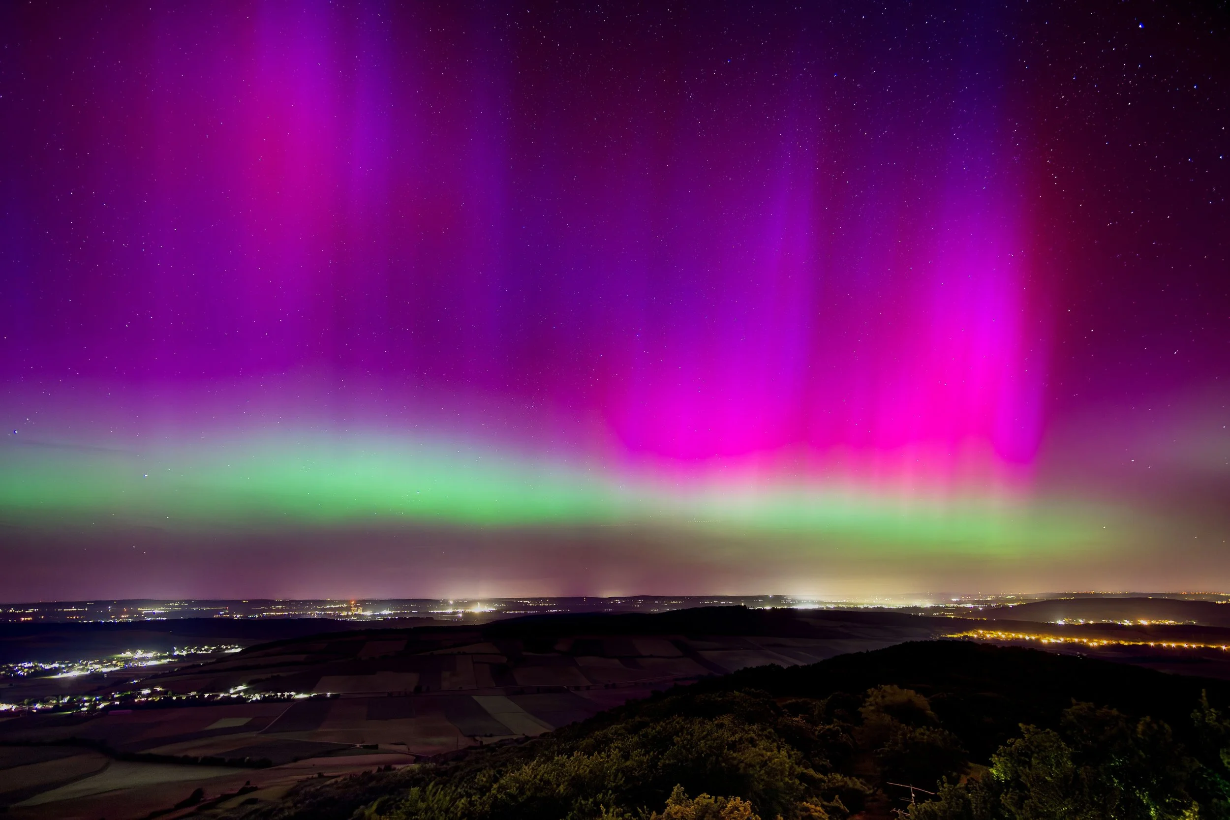 Nordlichter leuchten in Pink, Grün und Violett über eine nächtliche Landschaft mit Feldern und Bäumen.